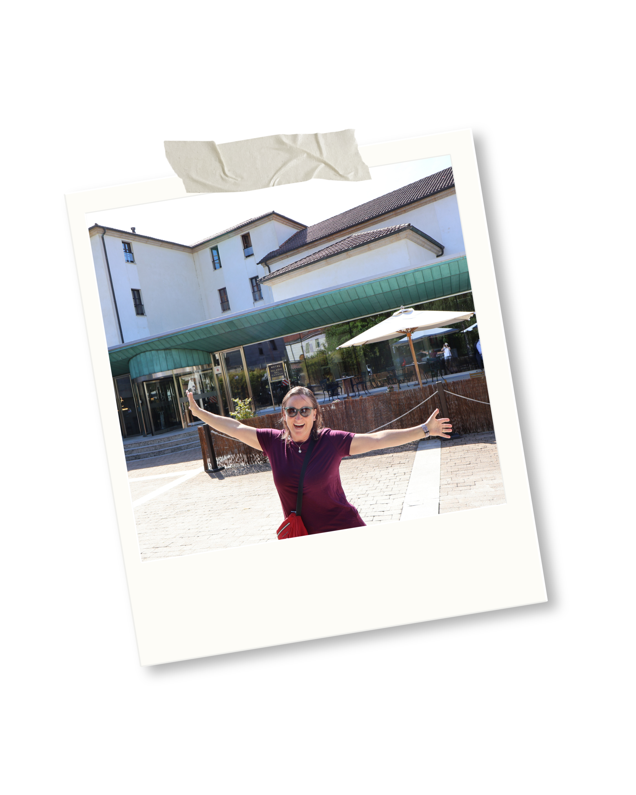 A woman with sunglasses and a maroon shirt with her arms outstretched, standing outside a modern building with a green awning and outdoor seating area.