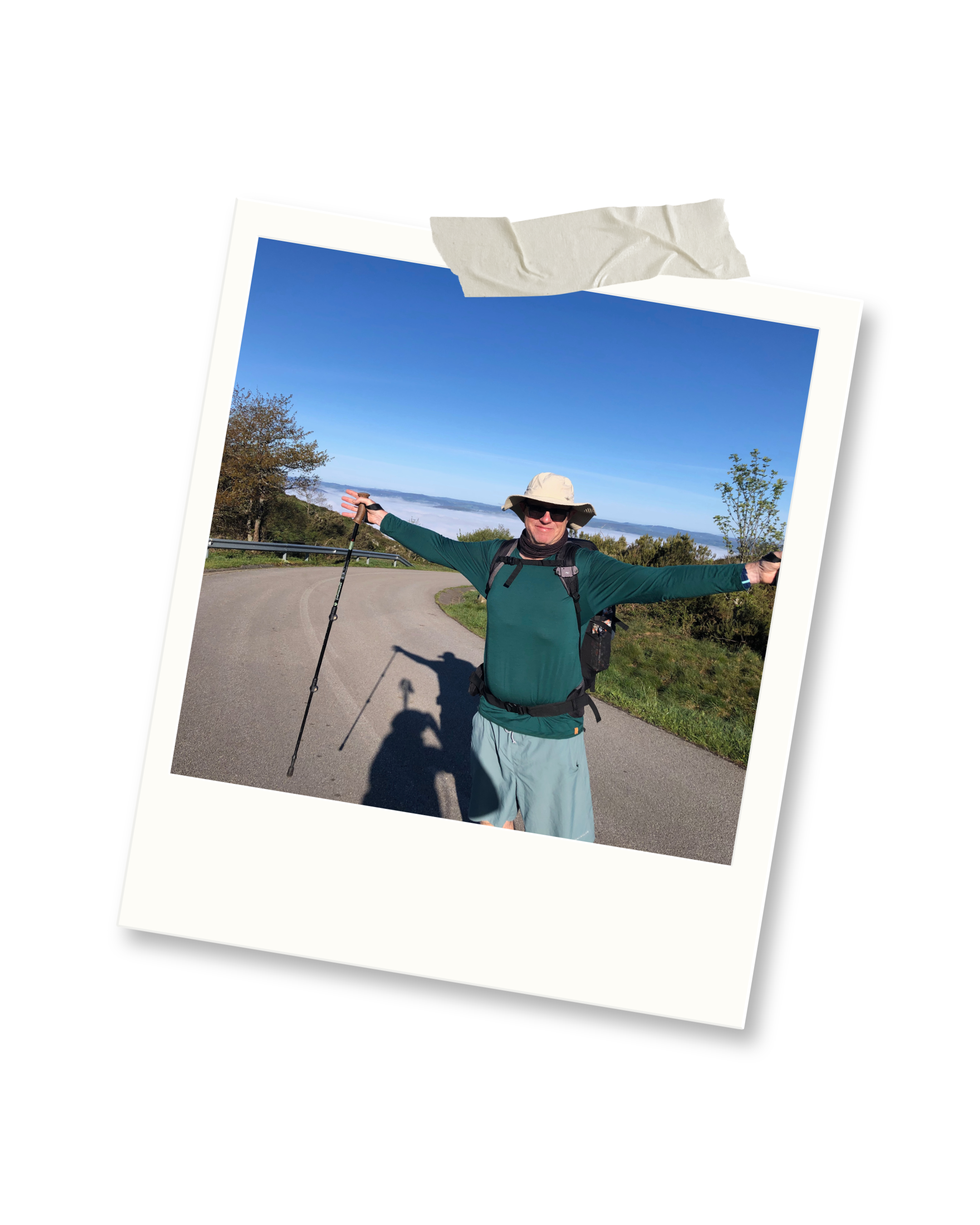 A man standing on a road with arms outstretched, holding a walking stick, wearing a sun hat, sunglasses, green long sleeve shirt, shorts, and backpack outdoors on a clear day with trees and a body of water in the background.