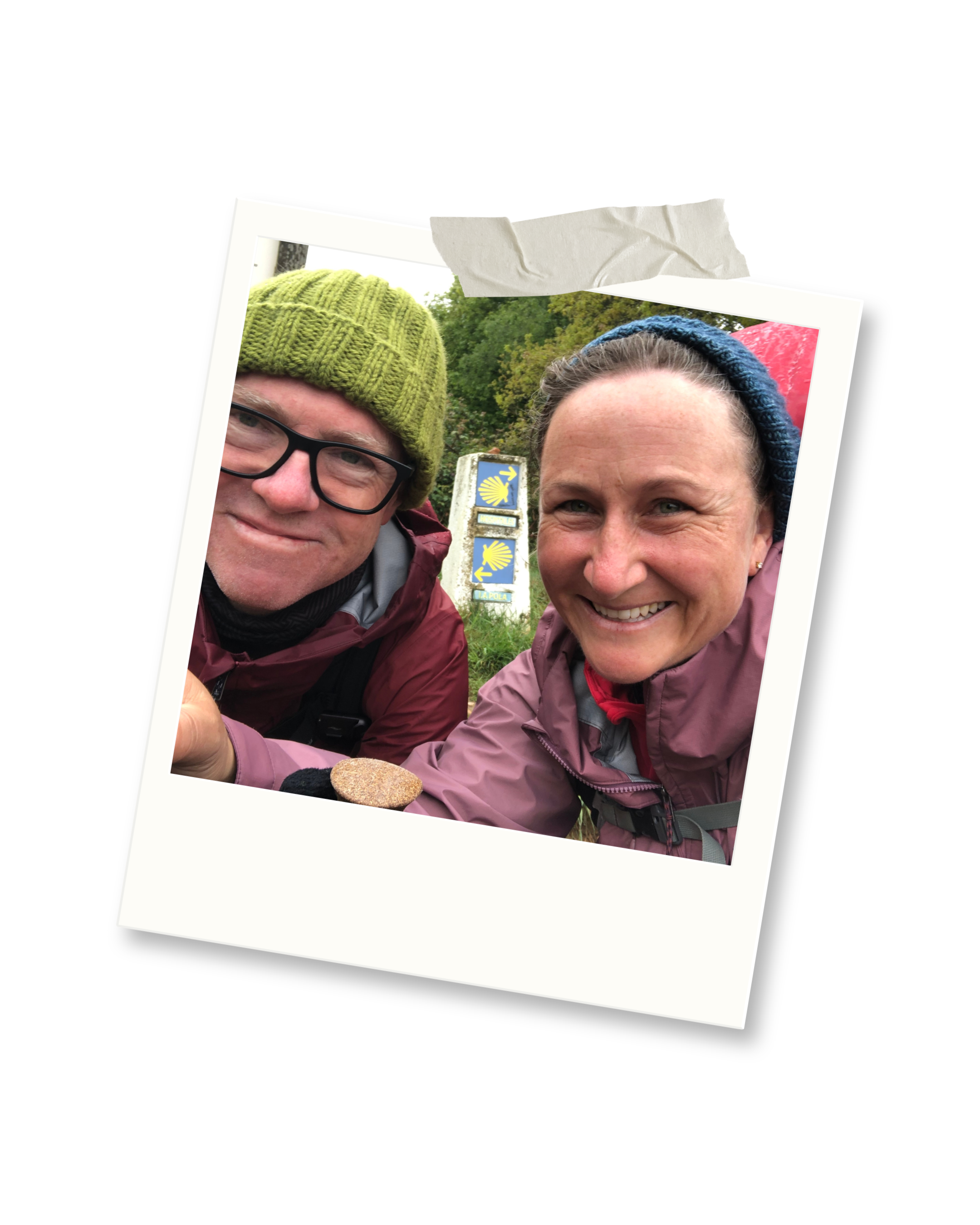 Two people smiling on a hiking trail, posing next to a signpost with a yellow shell symbol, indicating they are walking the Camino de Santiago, with greenery and trees in the background.