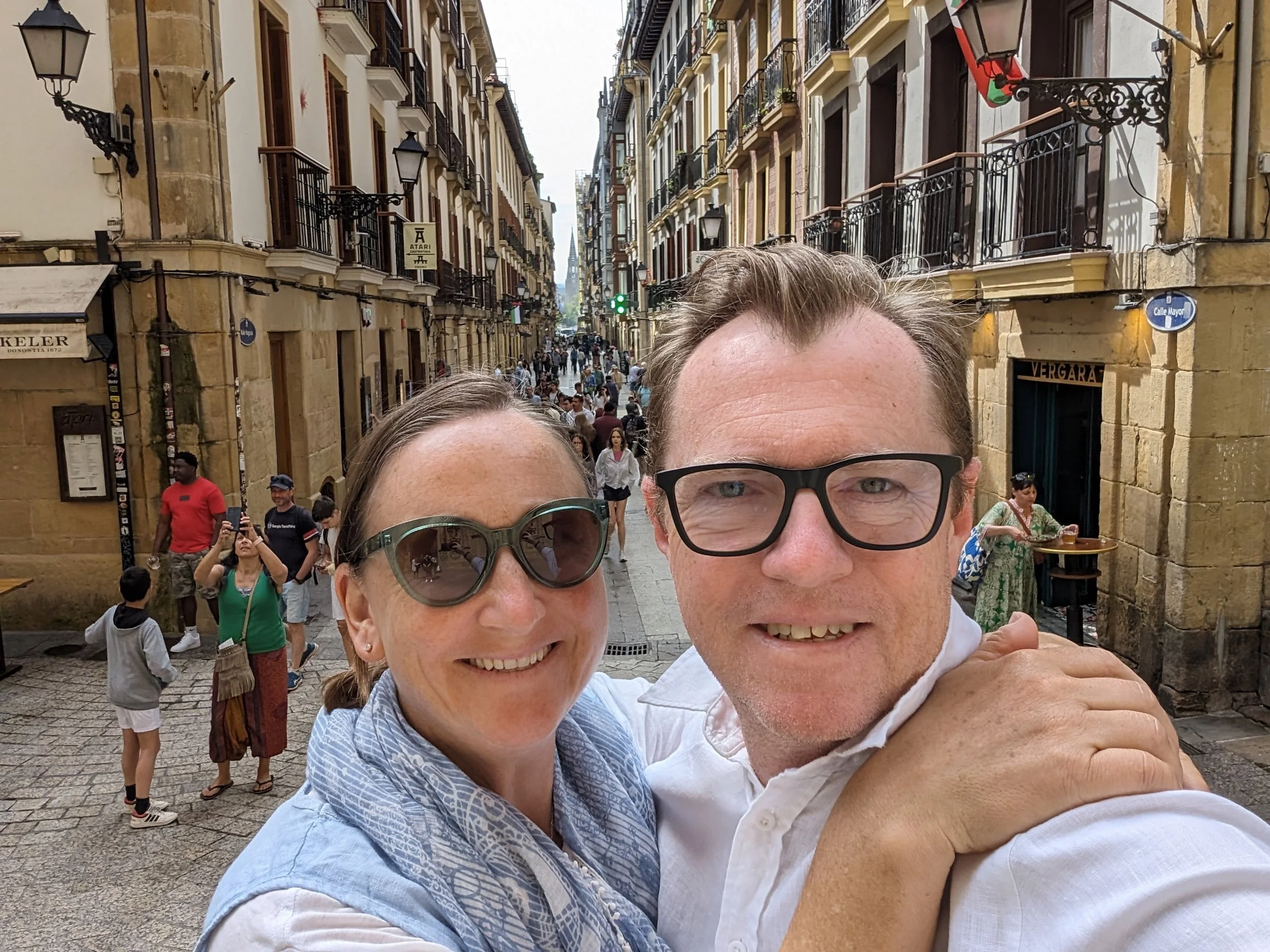Smiling couple taking a selfie on a busy street in an old European city, with stone buildings, balconies, and people walking.