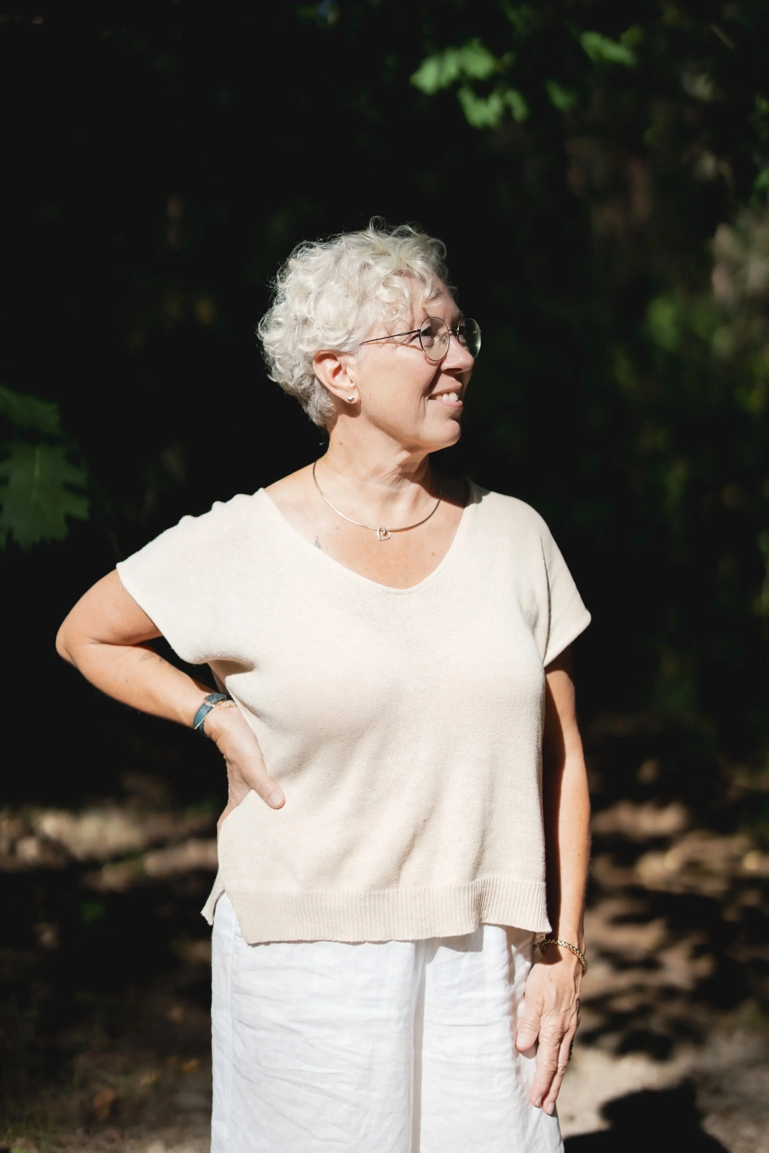 Een vrouw met grijs, krullend haar en brillen, die buiten staat, gekleed in een beige top en witte broek, met een hand op haar heup.