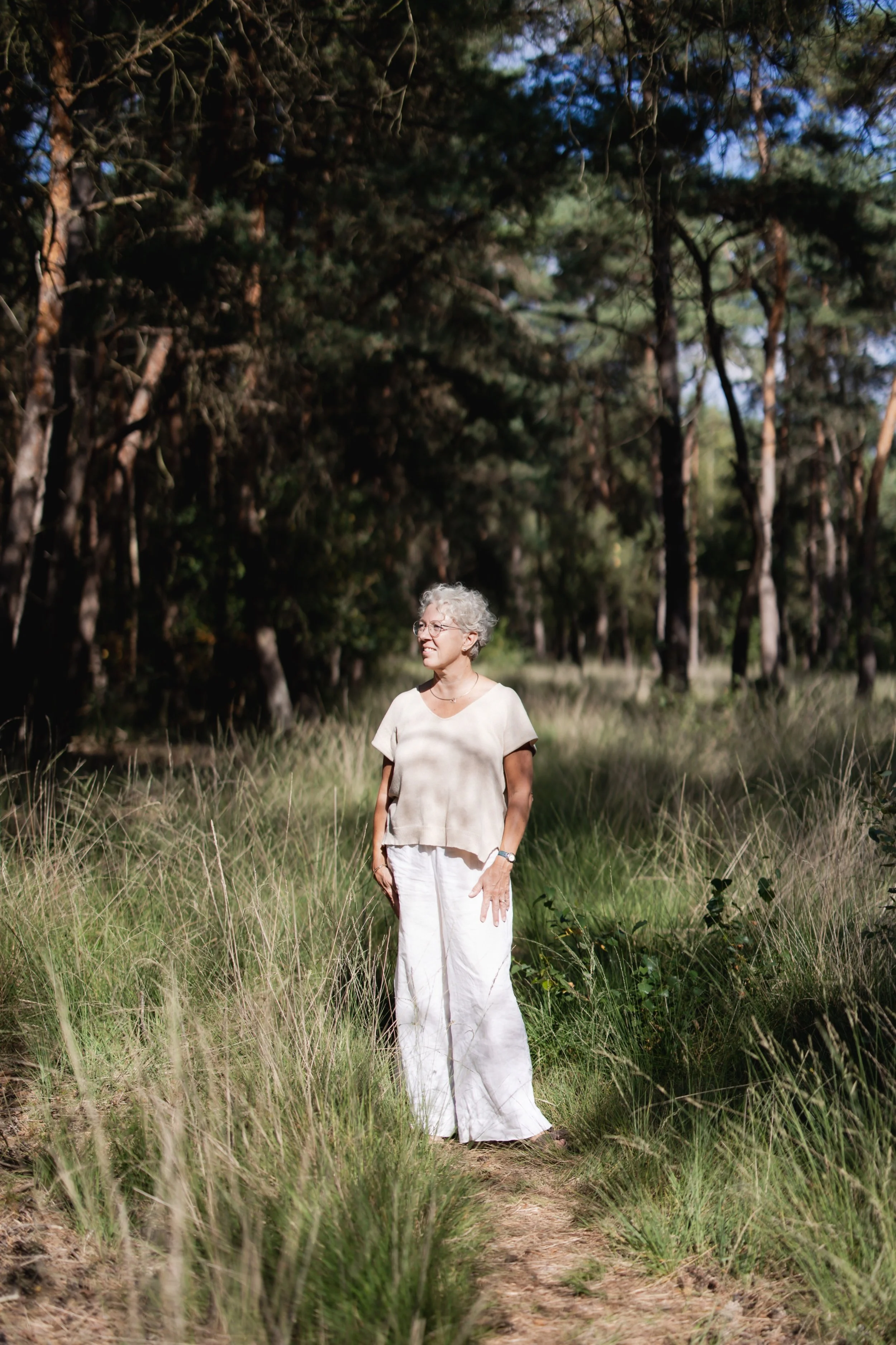 Oudere vrouw met witte haar en brillen, gekleed in een beige top en wijd uitlopende witte broek, staat op een pad in een bos, onder invloed van de zon en schaduw van de bomen.