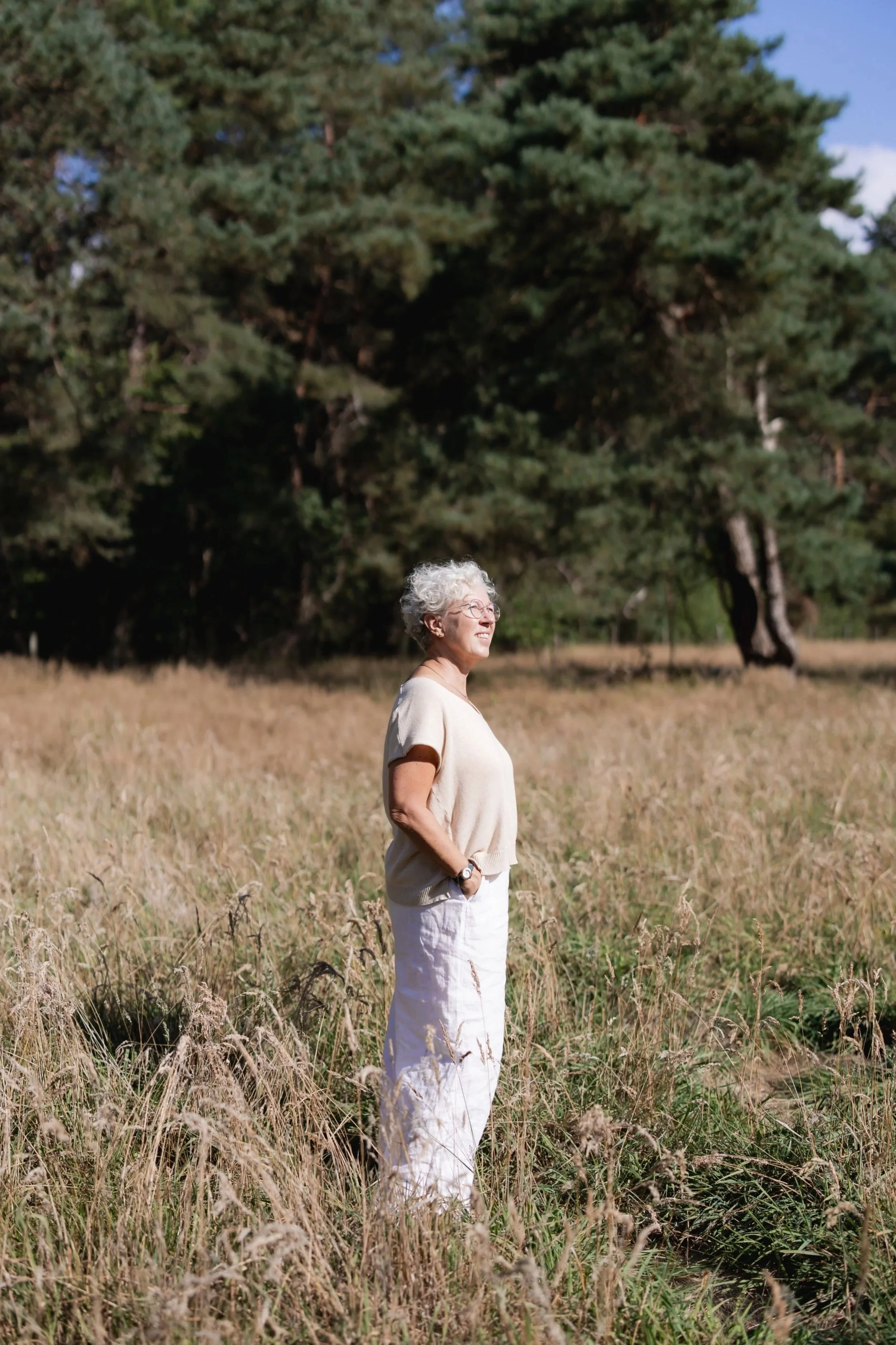Een oudere vrouw met wit krullend haar staat in een grasland met een grote dennenboom op de achtergrond, lachend in de zon.