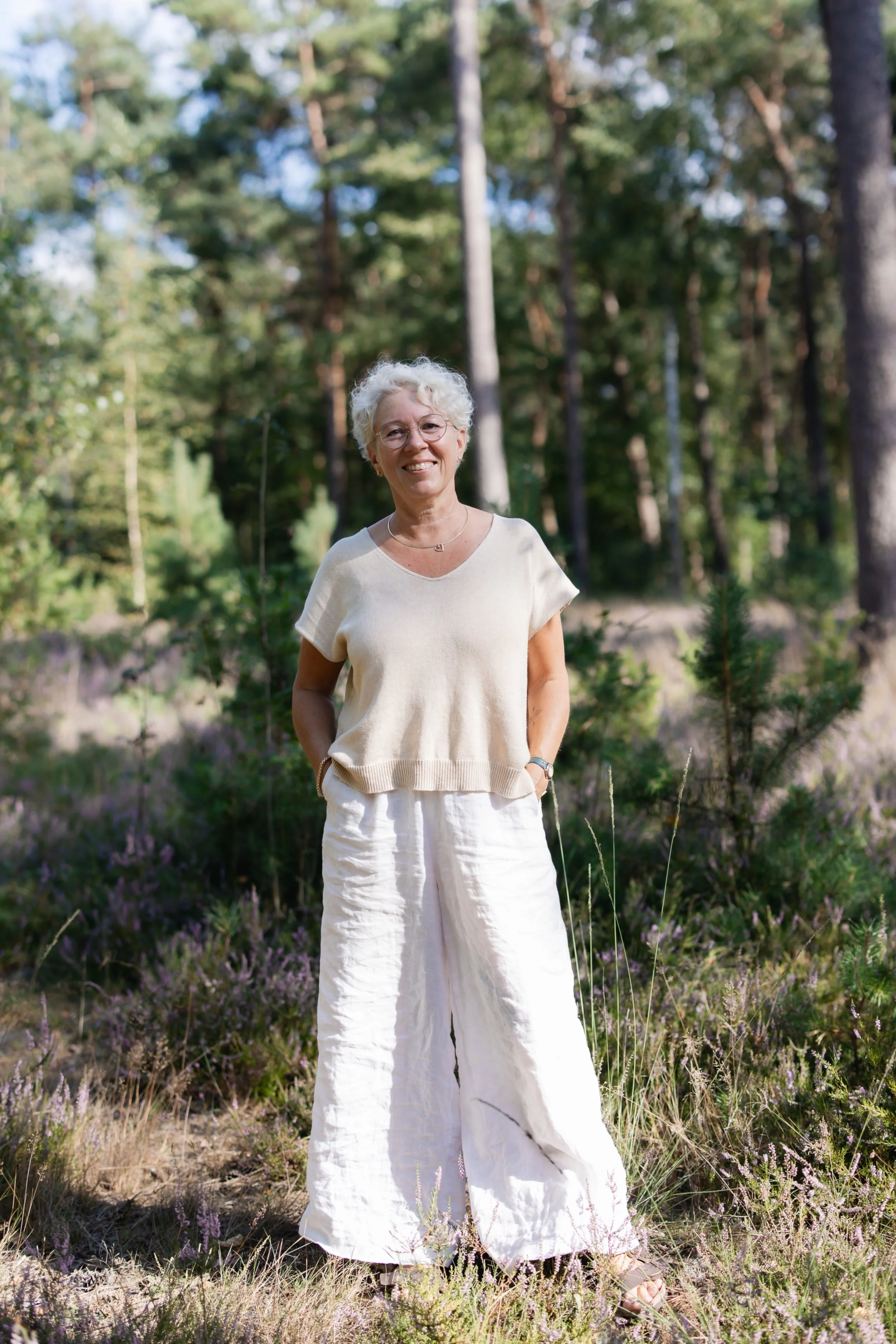 Een oudere vrouw met wit blond haar, brillen, een beige top en witte losse broek, staat in een bos, glimlacht.