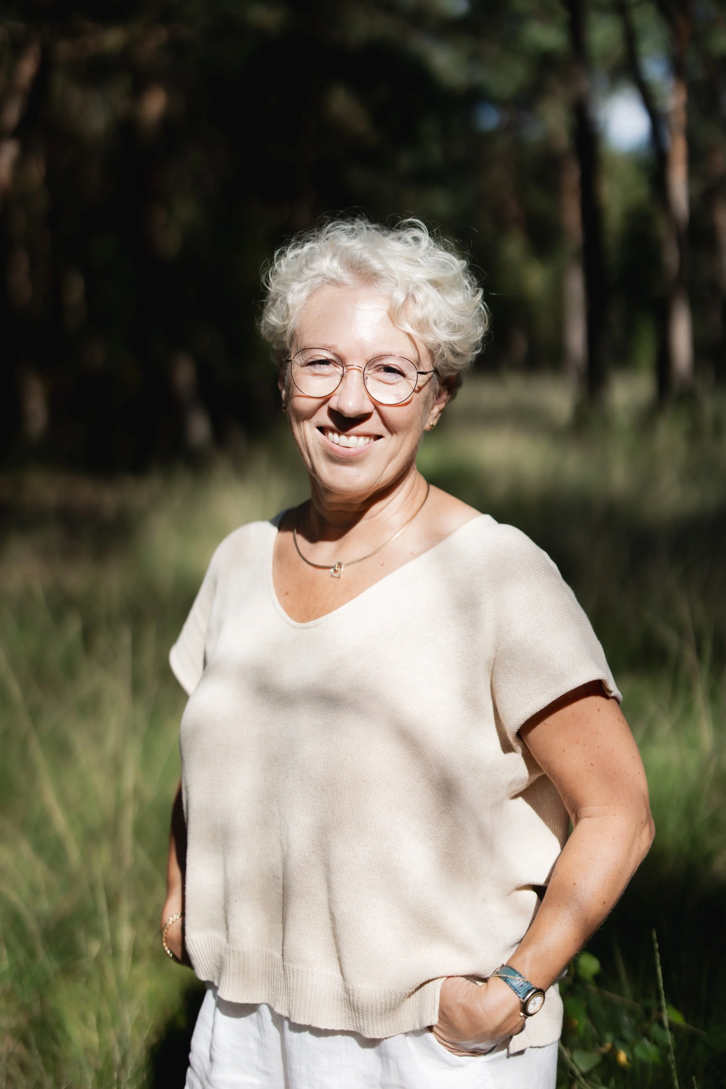 Een oudere vrouw met kort, krullend wit haar en ronde brillen staat buiten in een groene omgeving, glimlachend naar de camera.