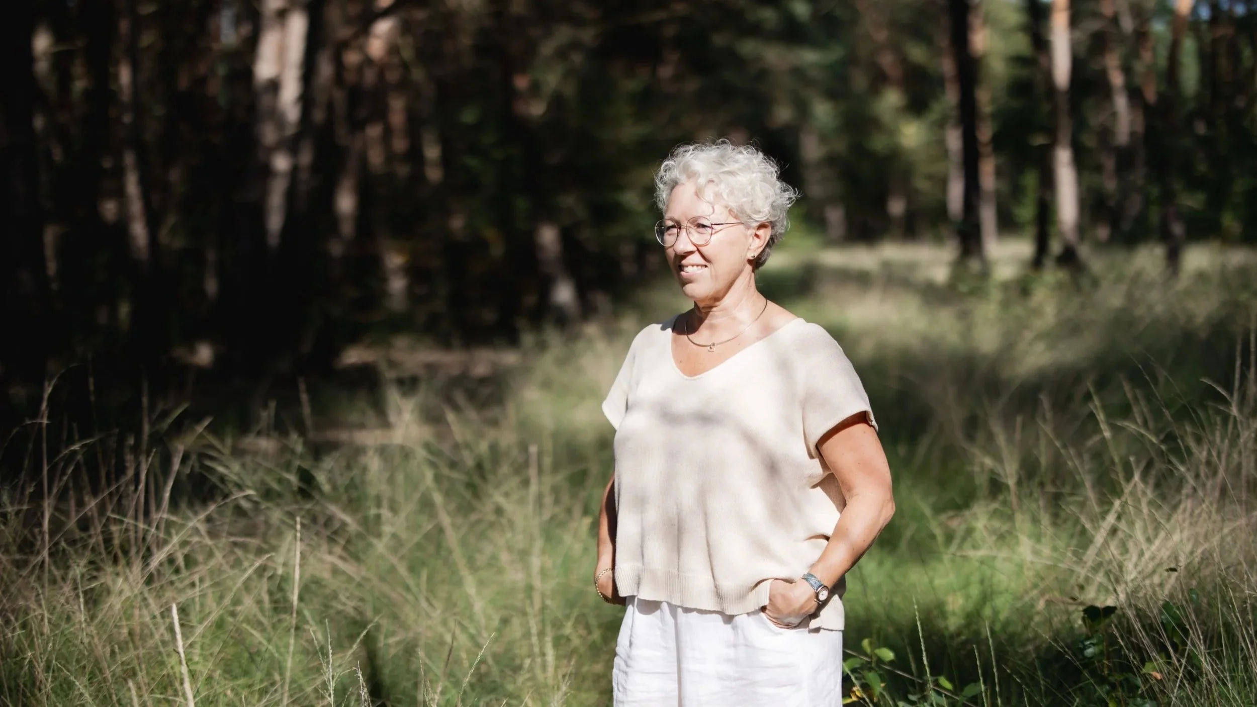 Een ouderwetse vrouw met grijs haar, bril, wit overhemd en wit rokje staat in een bos met bomen en gras, glimlachend naar de camera.
