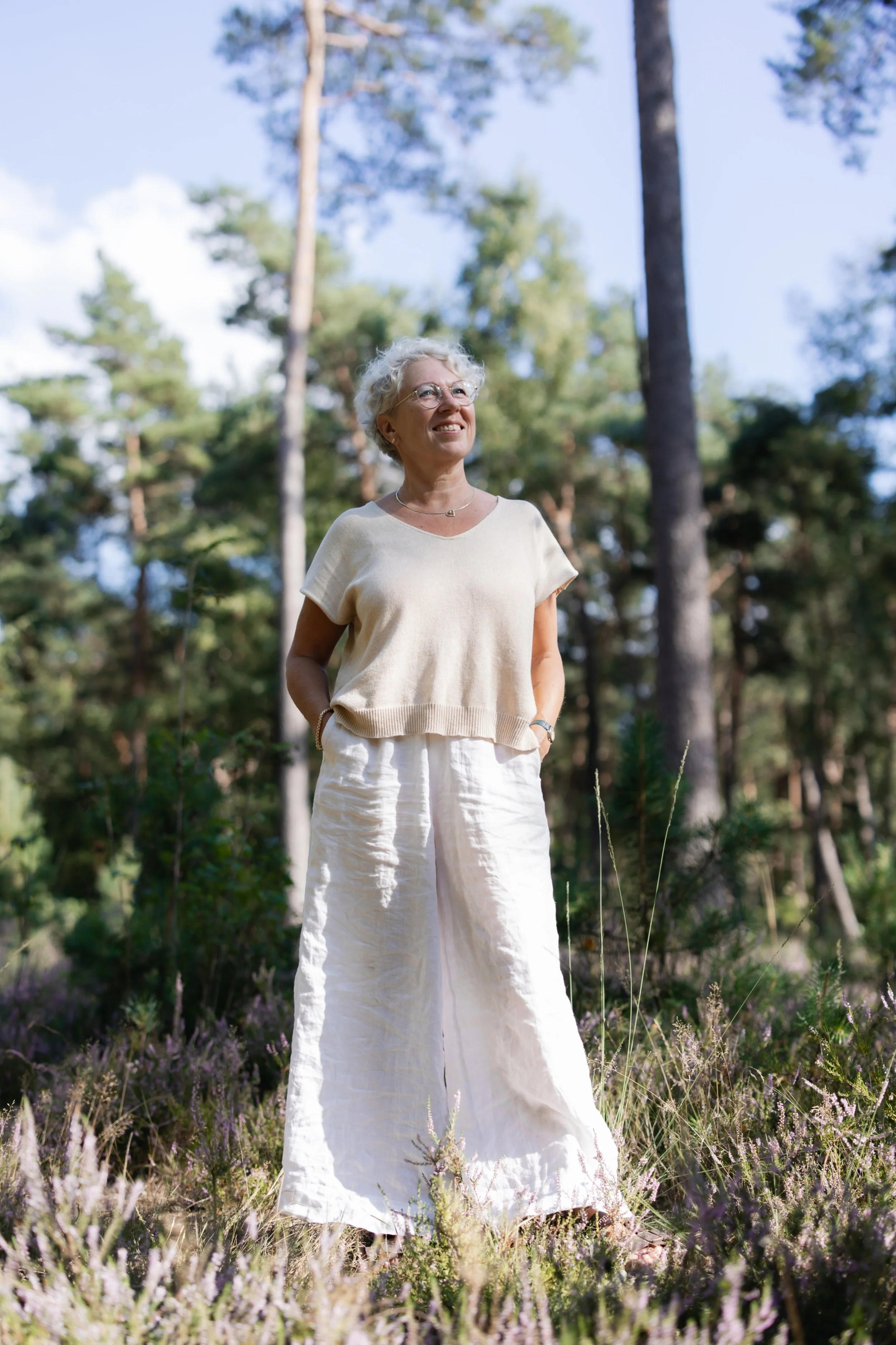 Een oudere vrouw met wit, krullend haar glimlacht terwijl ze in een bos staat, gekleed in een beige top en witte linnen broek.