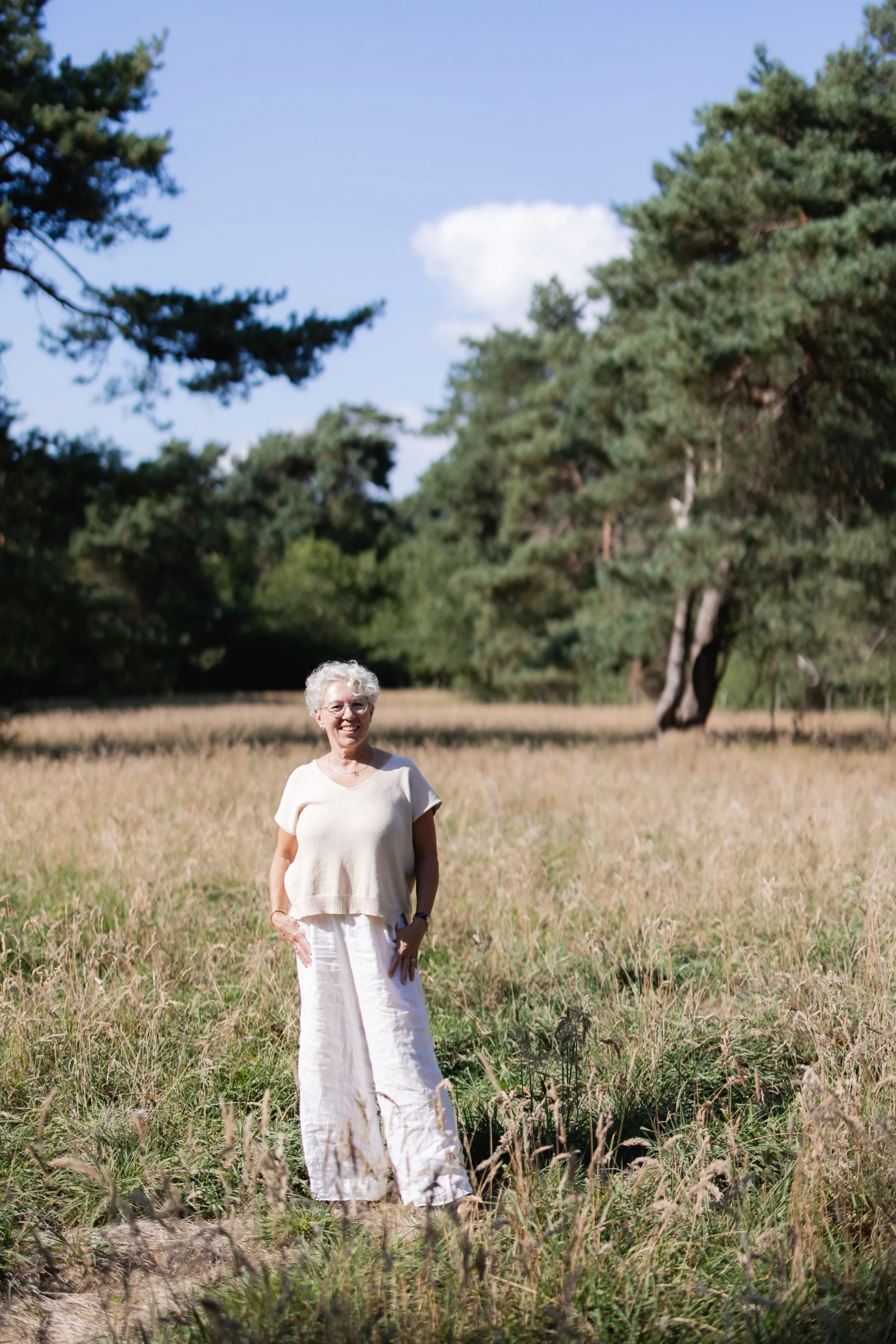 Vrouw met witte haar en witte kleding staat in een grasveld met bomen op de achtergrond op een zonnige dag.