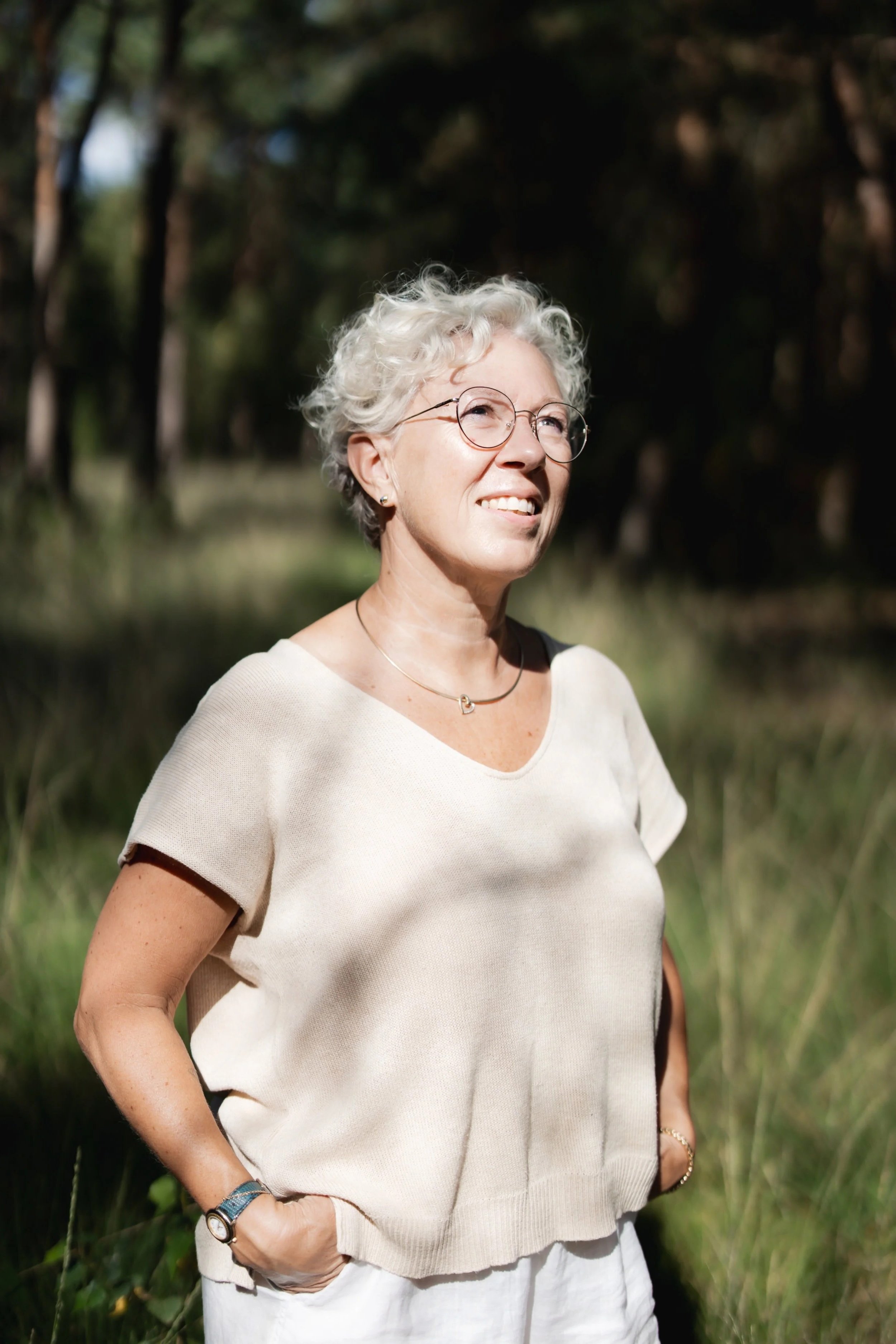 Een vrouw met grijze, krullende haar glimlacht terwijl ze buiten in een bos of park staat, gekleed in een lichte top en witte broek.