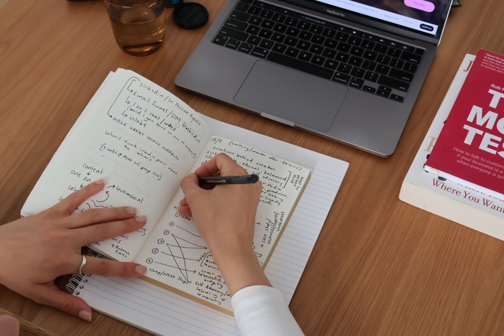 A person taking notes in a notebook at a wooden desk, with a laptop, a glass of water, and several books nearby.