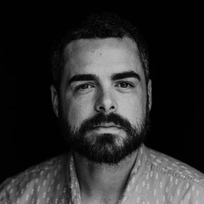 Black and white portrait of a man with dark hair and a beard, wearing a patterned shirt, looking directly at the camera.
