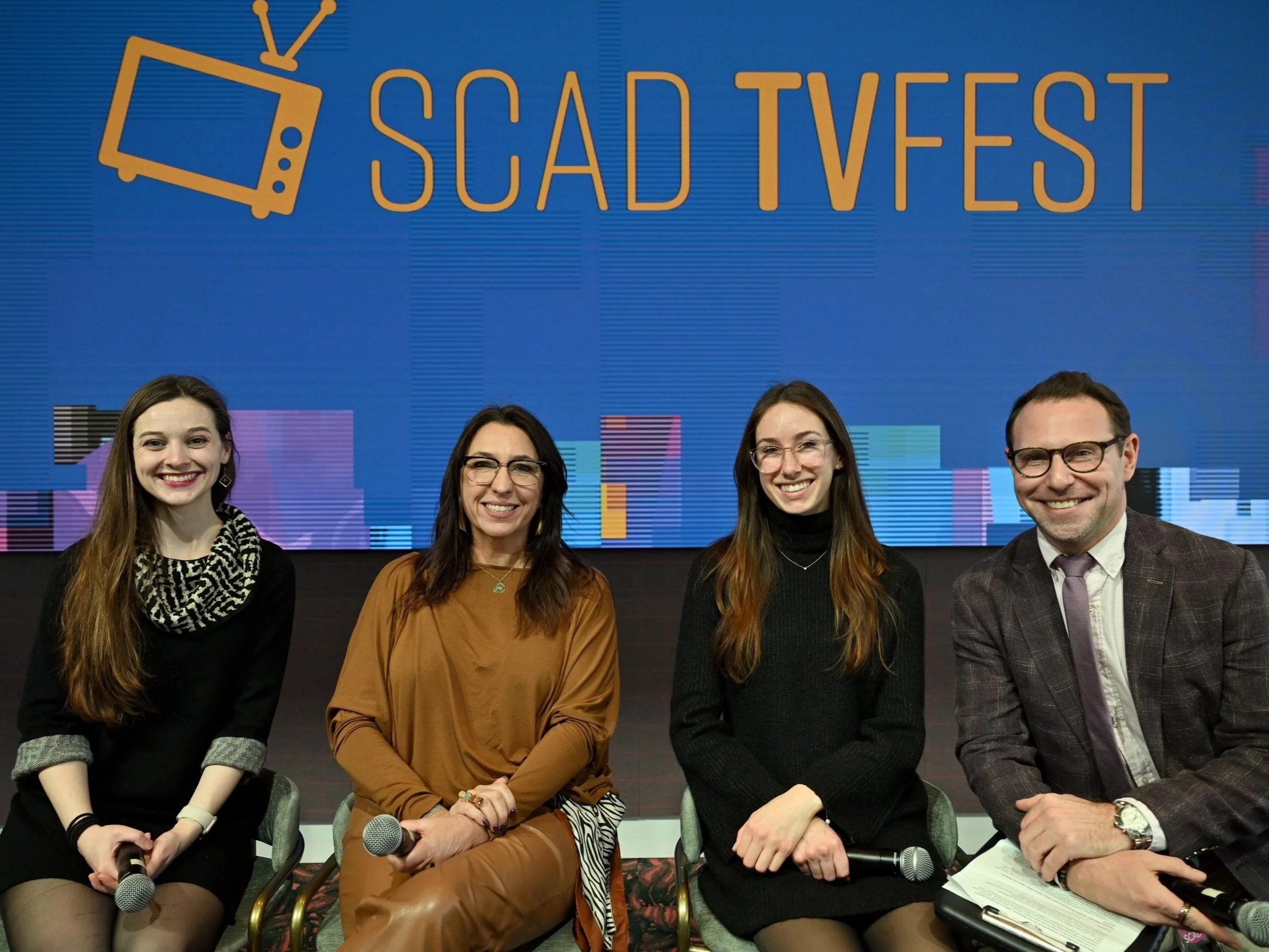 Four people sitting on stage at SCAD TV Fest, smiling, with a blue screen behind them displaying text 'SCAD TV FEST' and a TV icon.