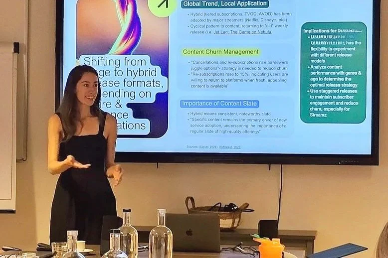 A woman in a black dress is giving a presentation in a conference room, standing next to a large screen displaying a slide on content management and trending topics.