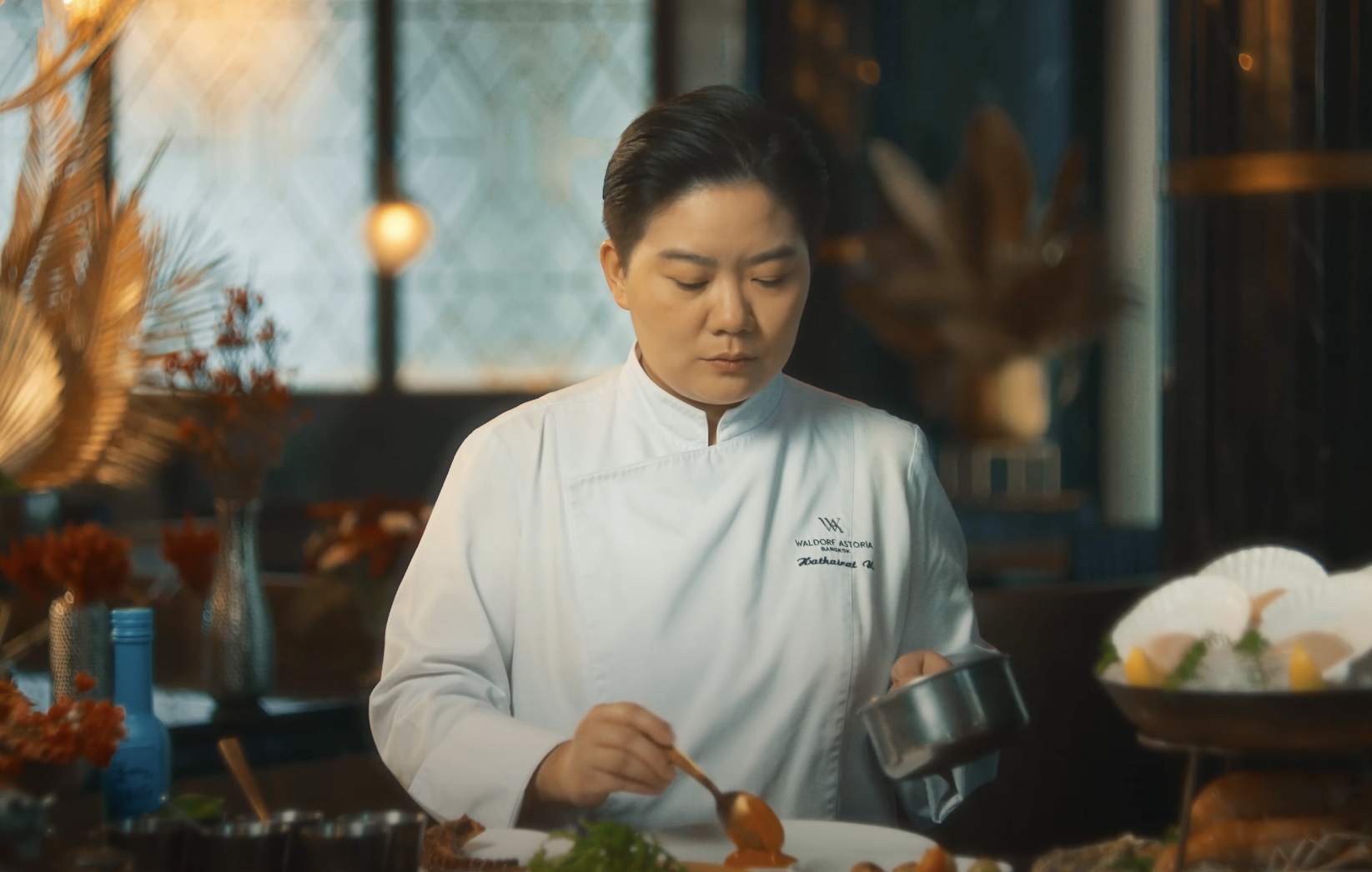 A female chef in a white uniform plating food in a restaurant kitchen with warm lighting and decorative elements in the background.