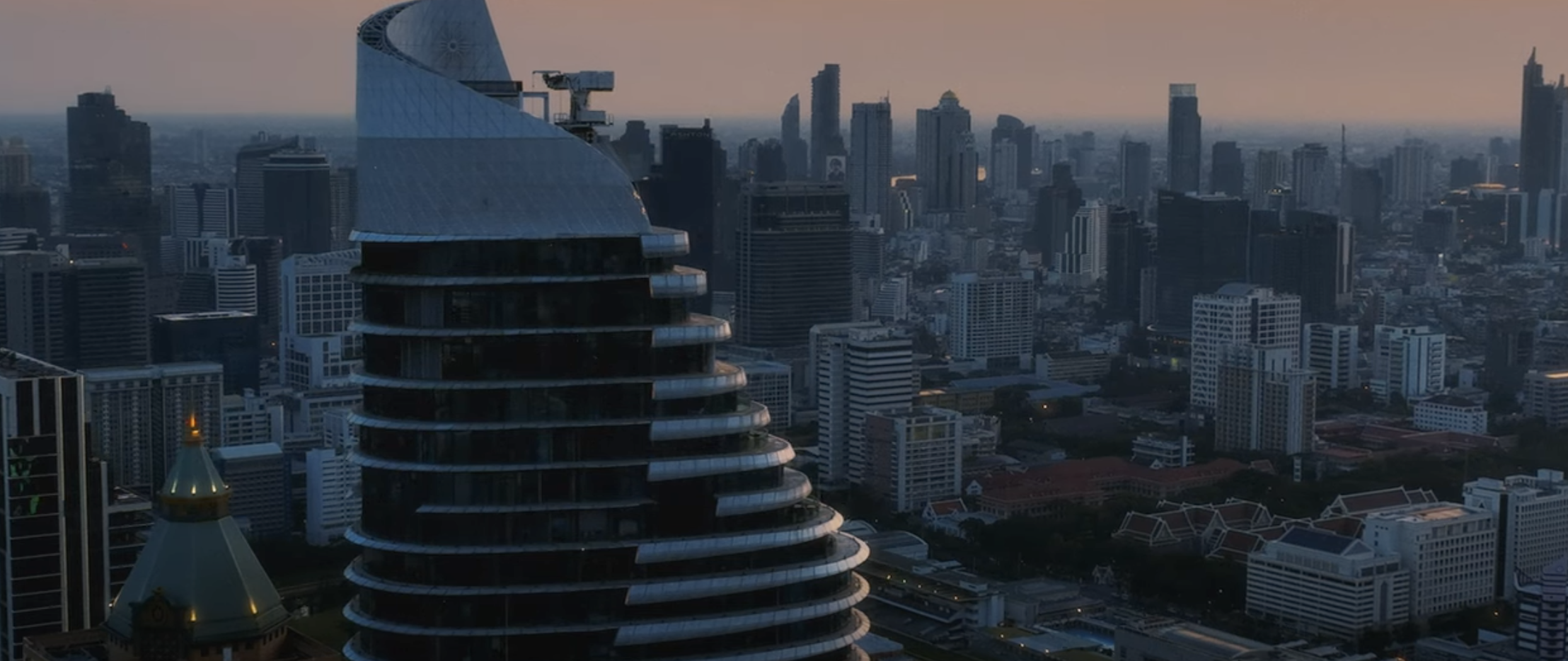 Skyline of a city with tall skyscrapers at dusk, featuring a prominent modern building with a rounded, stacked design in the foreground.