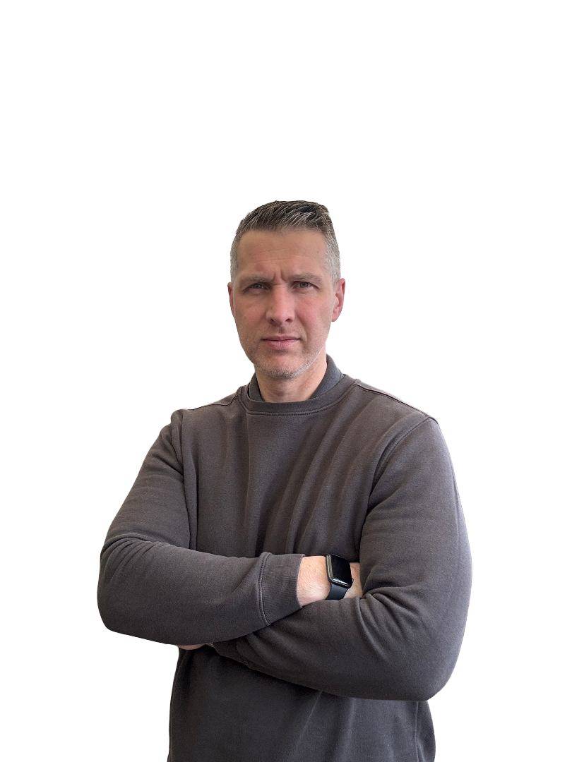 A man with short gray hair and a serious expression, wearing a dark gray sweatshirt and a smartwatch, standing with arms crossed against a plain background.