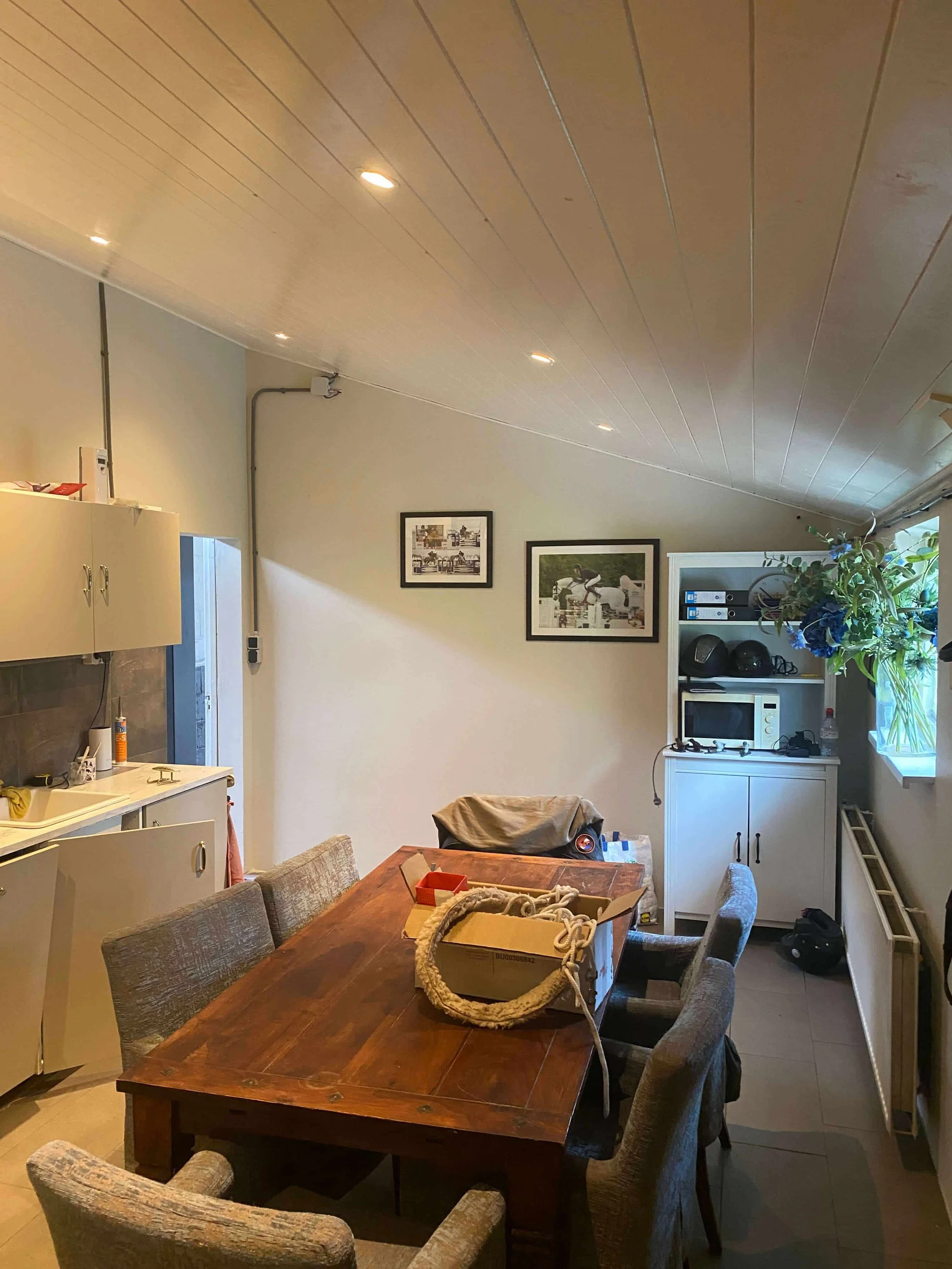 A cozy kitchen and dining area with a wooden table surrounded by upholstered chairs, a cabinet with appliances, and framed pictures on the wall.