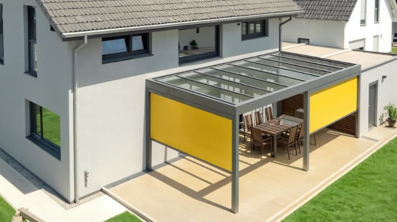 Moderne woning met een overdekt terras met houten tafel en stoelen, voorzien van een glazen dak en felgele windschermen.