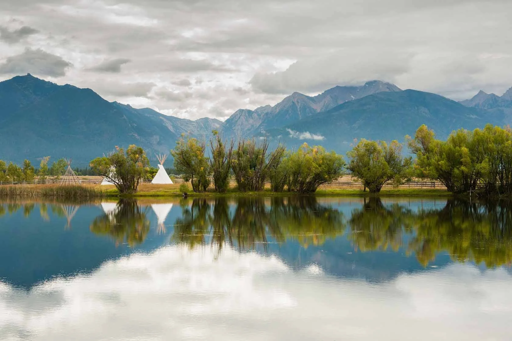 A scenic landscape with a calm lake reflecting green trees and a cloudy sky, mountains in the background, and a white teepee near the water's edge.