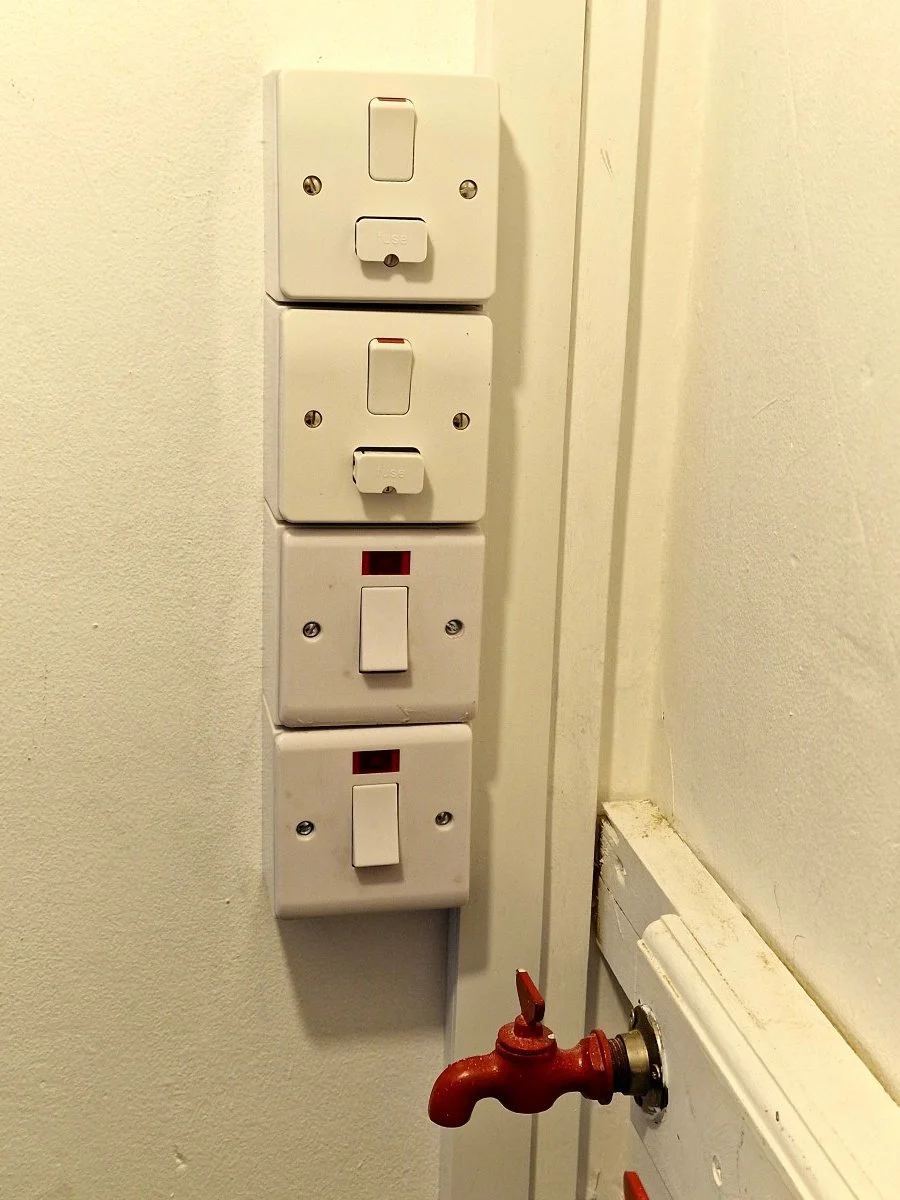 A wall with three switches and two electrical outlets above a red water valve.