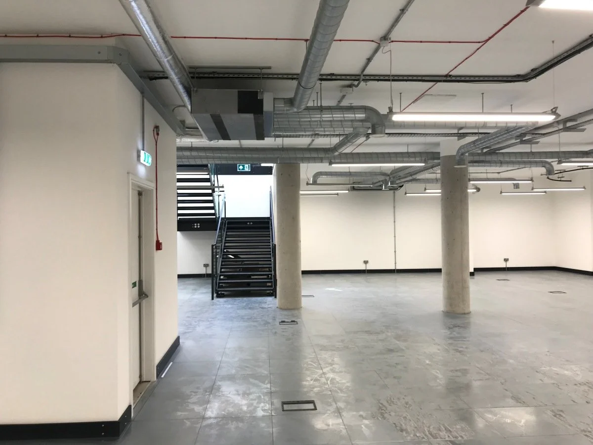 New empty indoor commercial space with grey tiled floor, concrete support columns, exposed ductwork and pipes on the ceiling, a staircase at the back leading to an upper level, and white walls with a new fire alarm and electrical installation  