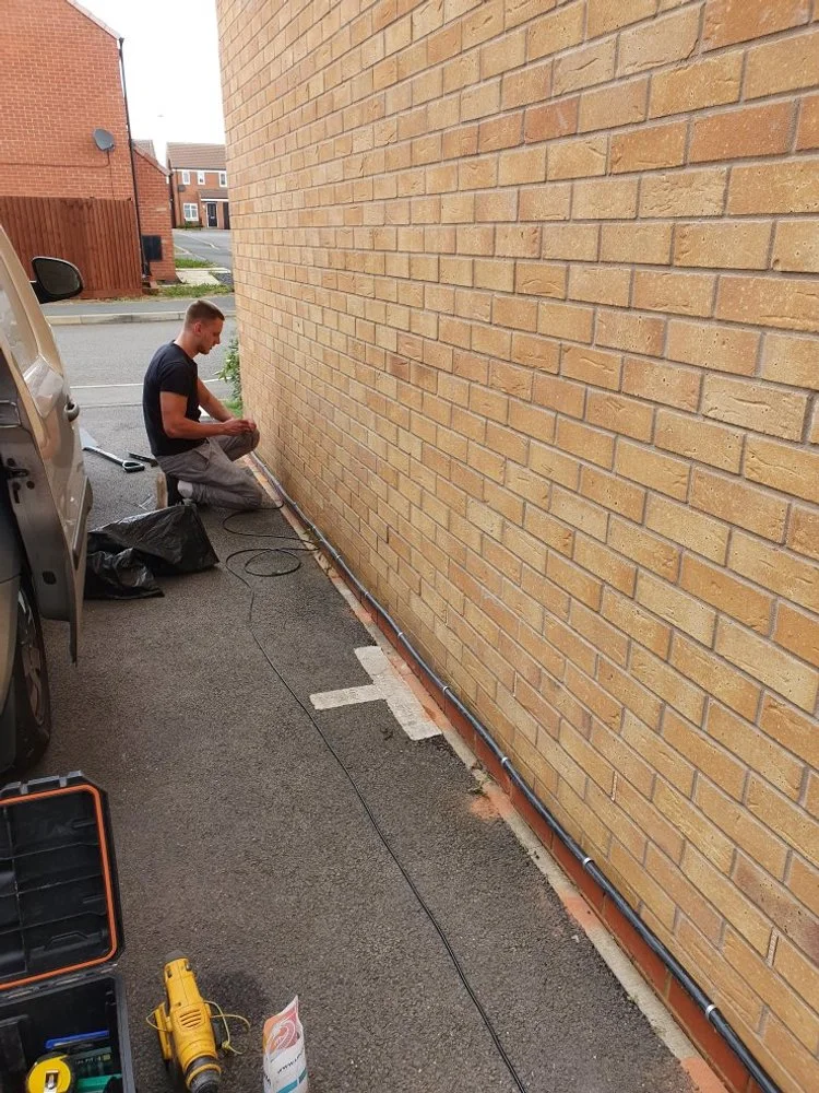 A man is working on electrical wiring along the base of a brick wall outside a building, with tools and equipment nearby on the ground.