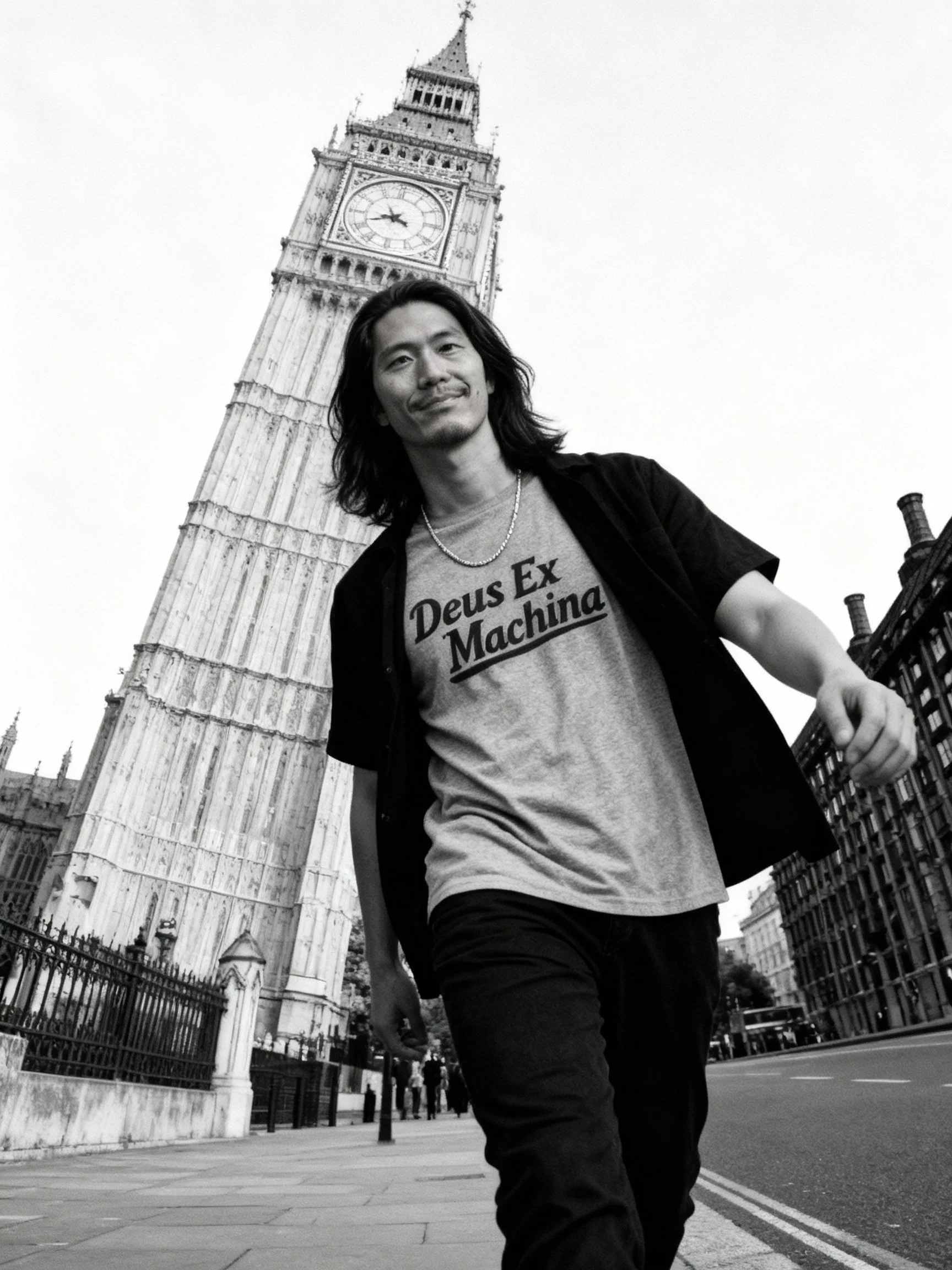 A man with long hair smiling and walking on the street in front of Big Ben in London, wearing a t-shirt that says 'Deus Ex Machina' and an open shirt over it.