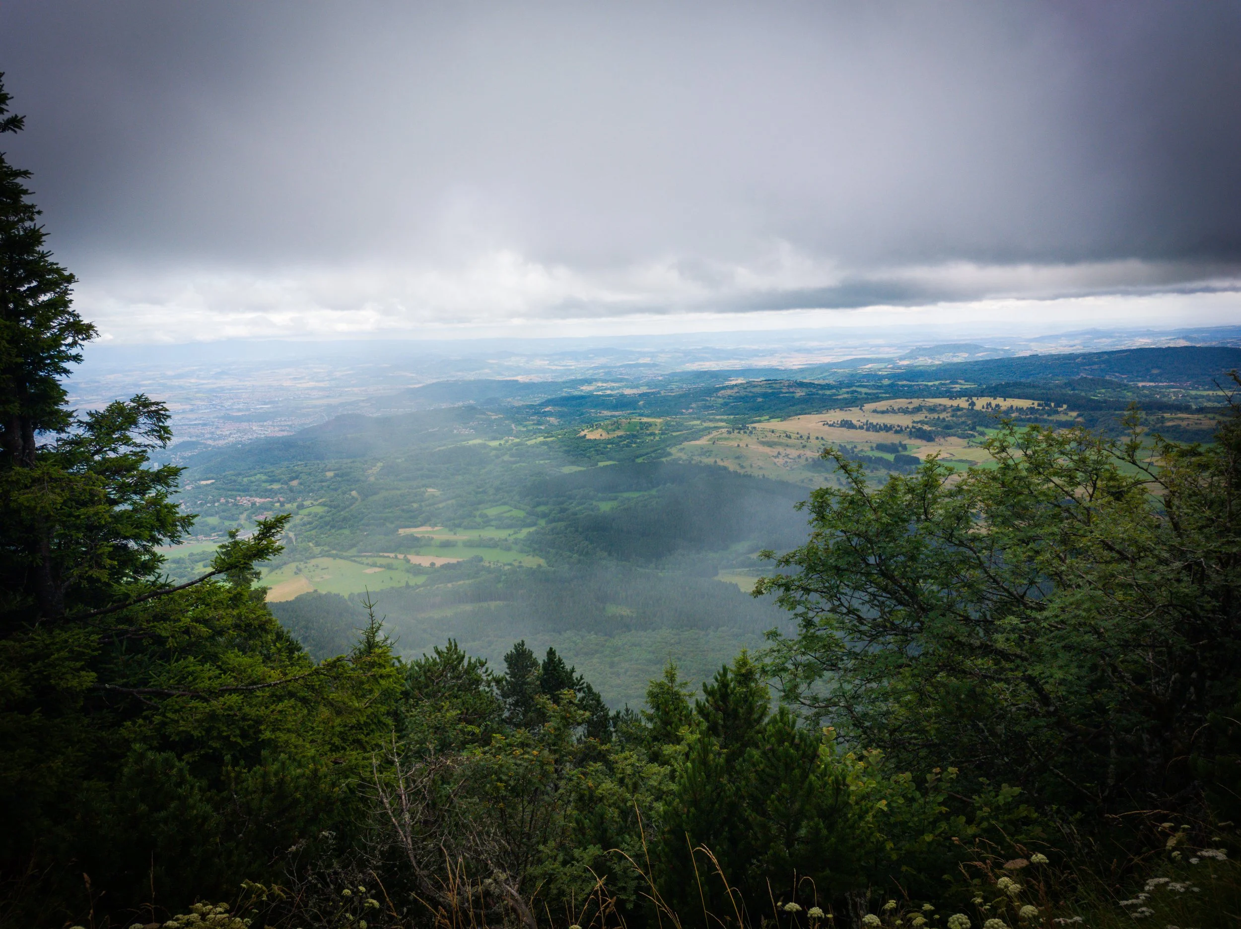 View of a lush, green valley with trees in the foreground, hills and fields in the distance under a dark, cloudy sky.