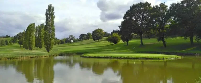 Golf course with water hazard, green grass, trees, and cloudy sky.