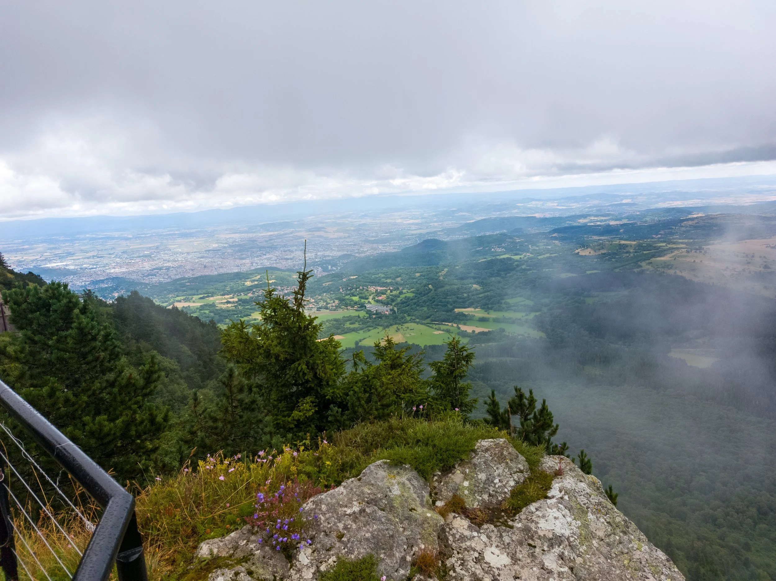 View from a mountain lookout showing green forests, fields, and a city in the distance under a cloudy sky with some mist and rocks and plants in the foreground.