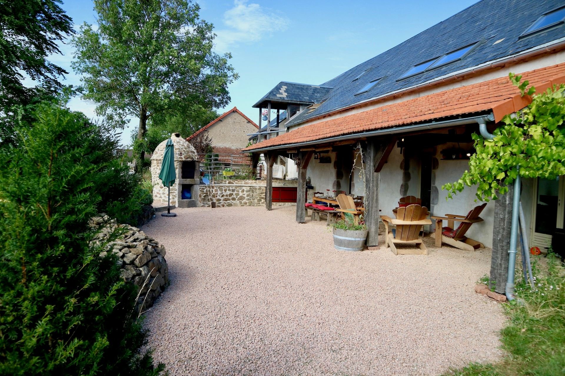 A spacious outdoor patio area of a rustic house with wooden supports, Adirondack chairs, a barrel planter, and a gravel ground. There is also a stone oven or pizza oven and lush green trees surrounds the area.