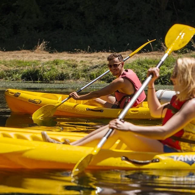 Two people kayaking on a calm river, one man with sunglasses and short hair in a red life vest, and a woman with long blonde hair in a red and black life vest, both paddling yellow kayaks.