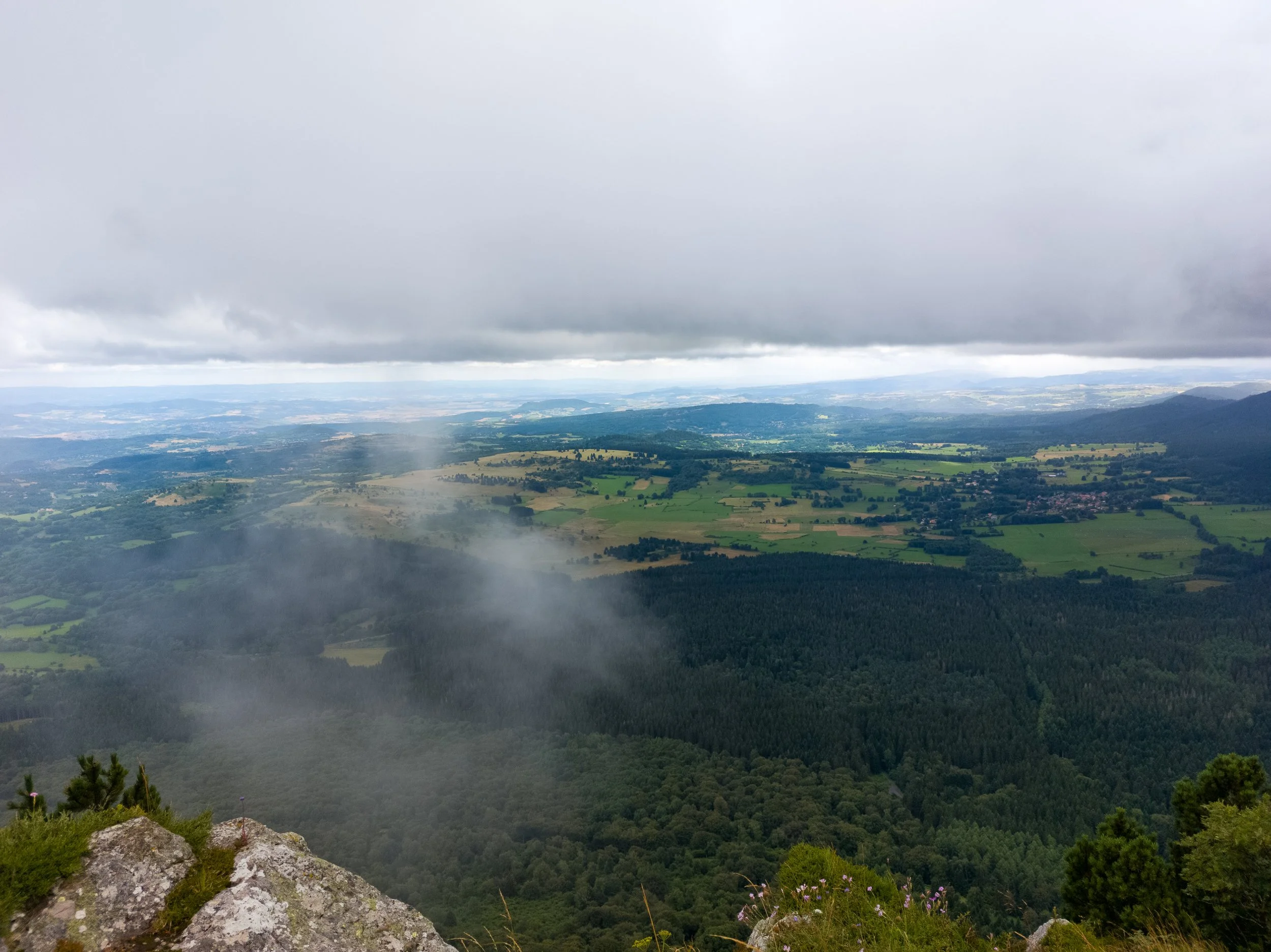A scenic view from a mountain overlooking a rural landscape with farmlands, forests, and scattered houses under a cloudy sky.