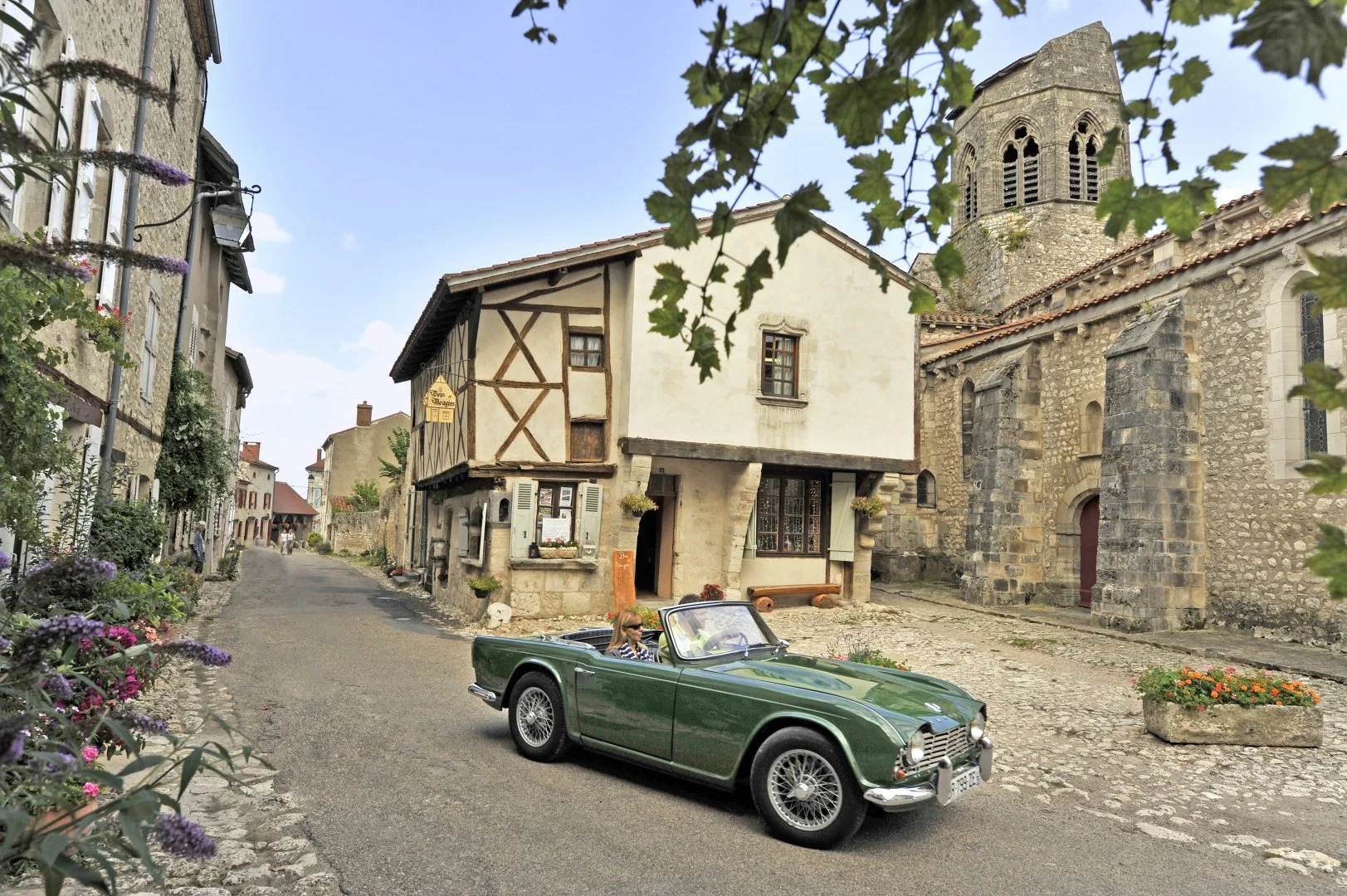 A vintage green convertible car drives through a historic European village with cobblestone streets, stone and timber houses, and a stone church tower in the background. A woman is driving, and the sky is partly cloudy with trees and flowers lining the streets.
