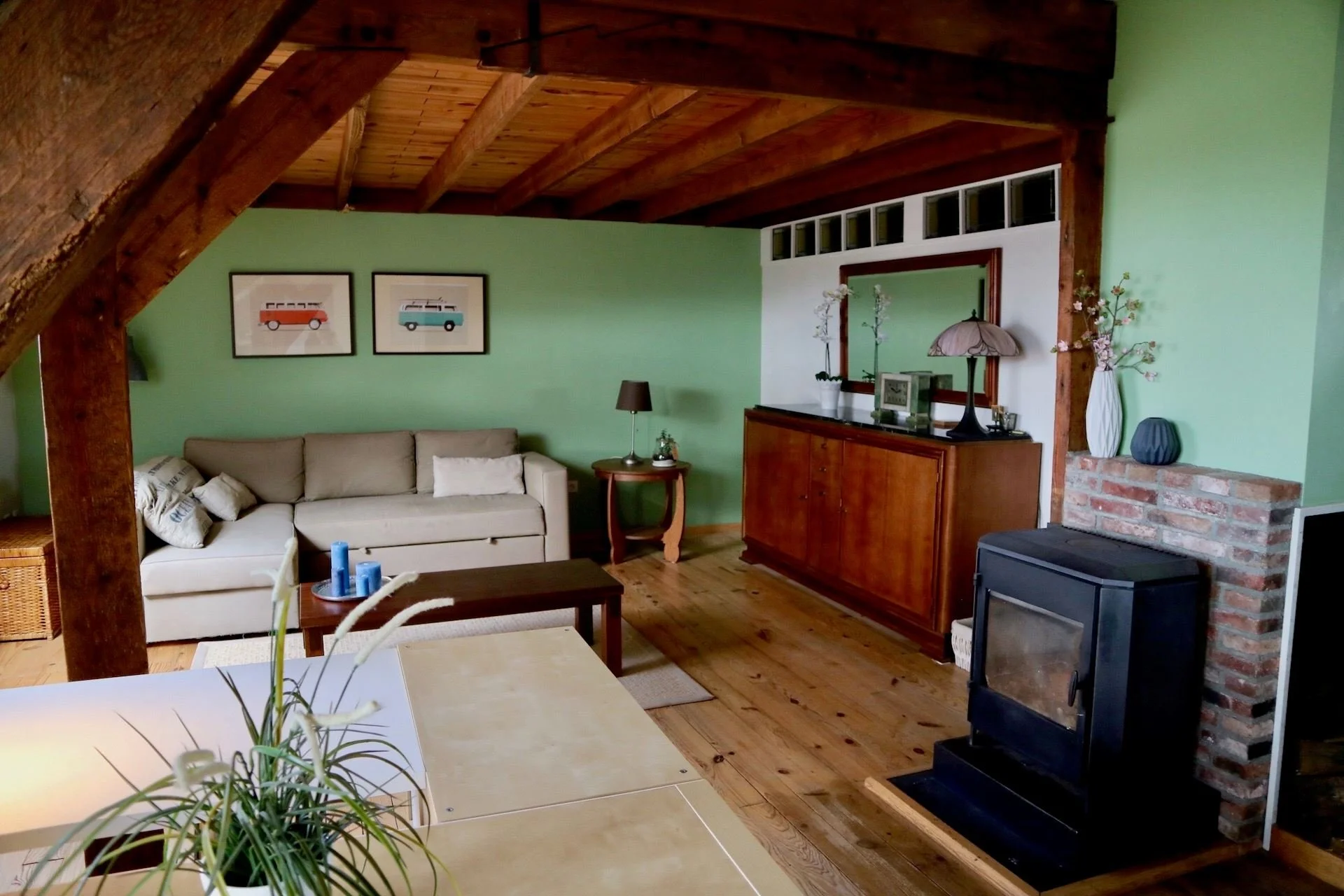 Living room with light green walls, wooden ceiling, beige sofa, wooden coffee table, framed art of colorful vintage vans, wooden sideboard with decorative lamp and vases, fireplace, and various decorative items.
