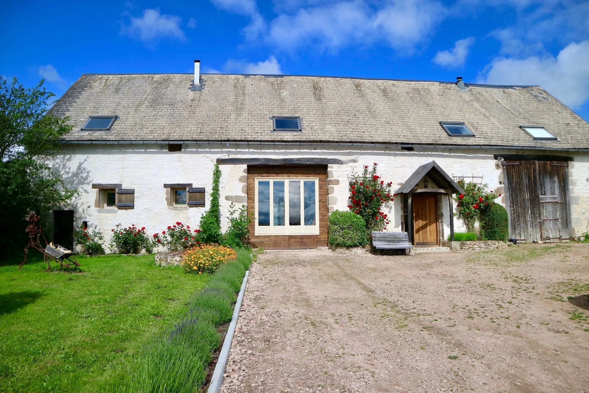A charming white cottage with a slate roof, surrounded by colorful flowers and greenery, with a gravel driveway and a wooden bench outside under a blue sky with clouds.