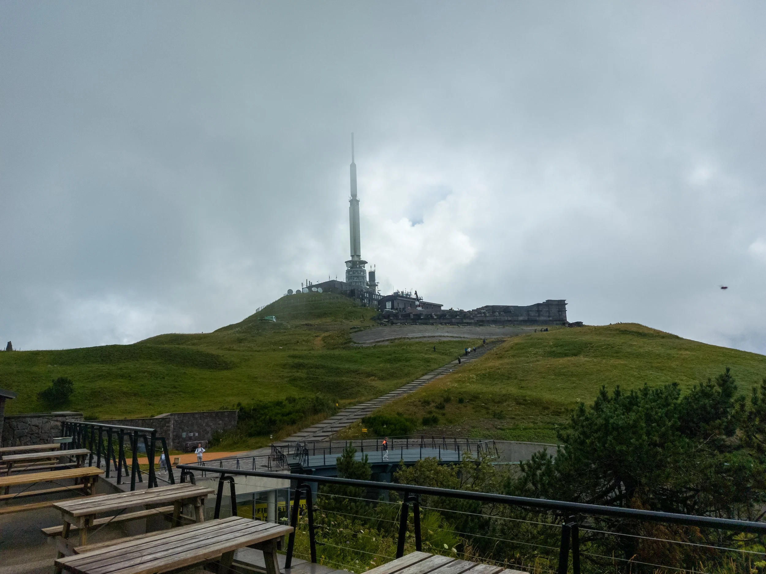 A hilltop with a large antenna tower and a building, overlooking a grassy area with some trees. The foreground has outdoor seating with benches and a railing. The sky is cloudy.