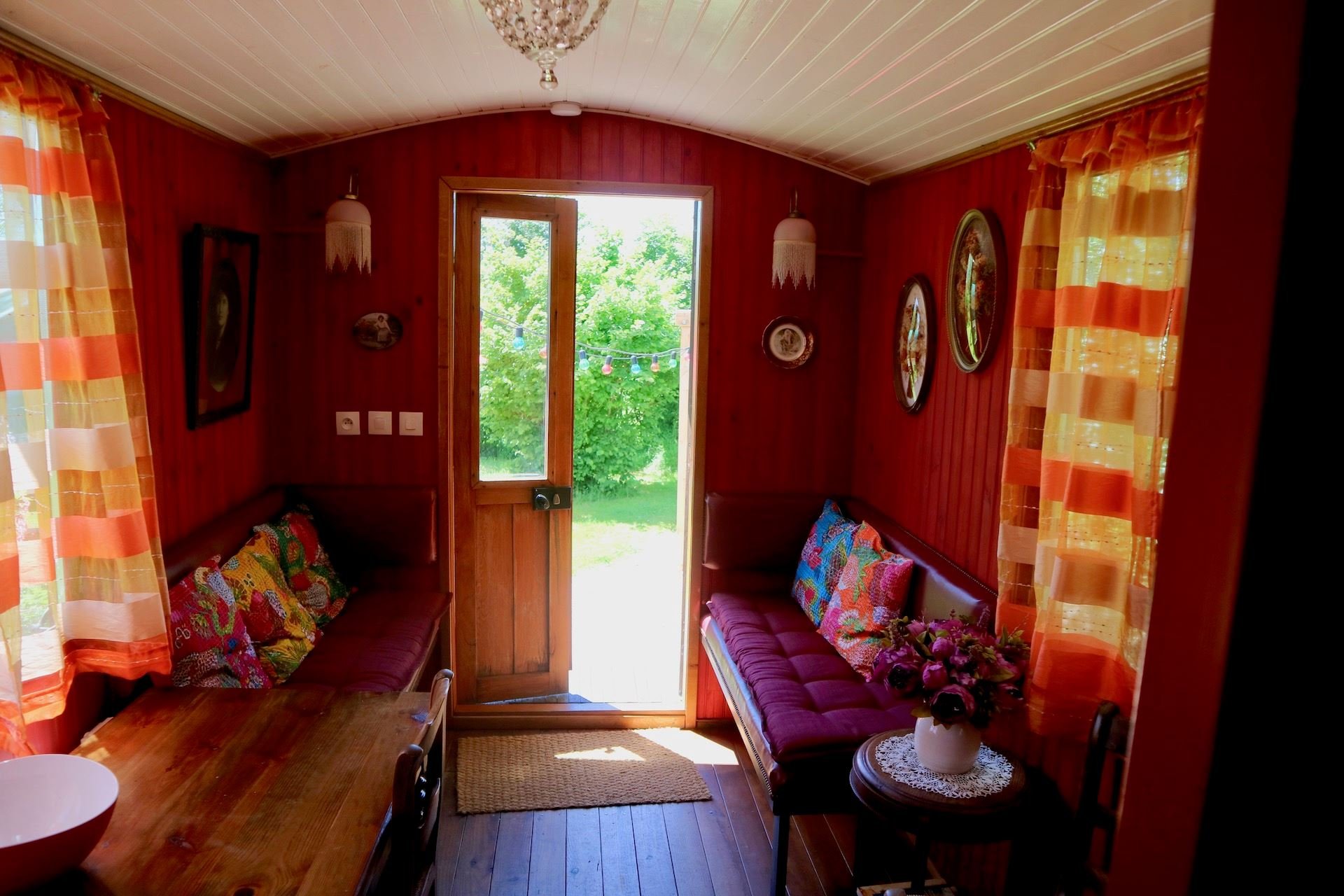 A cozy porch interior with red wooden panel walls, orange curtains, two benches with colorful cushions, a wooden table, a vase of flowers, and a door leading outside to a garden with green trees and string lights.