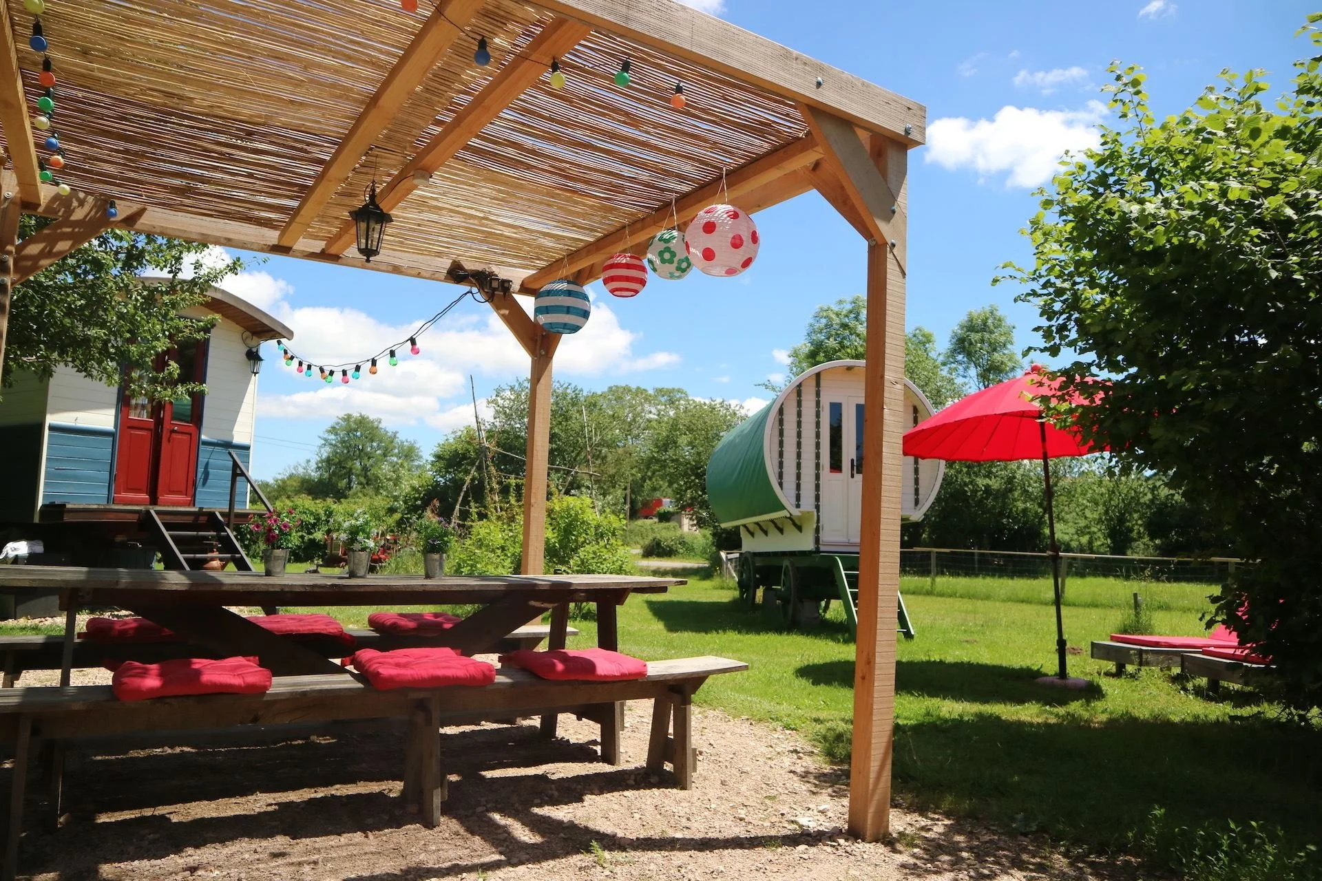 An outdoor backyard scene with a wooden pergola decorated with hanging colorful paper lanterns and strings of multicolored lights. There is a picnic table with red cushions underneath the pergola, and a red umbrella with a matching lounge chair nearby. In the background, a tiny green and white house on wheels and lush green trees are visible under a blue sky with scattered clouds.