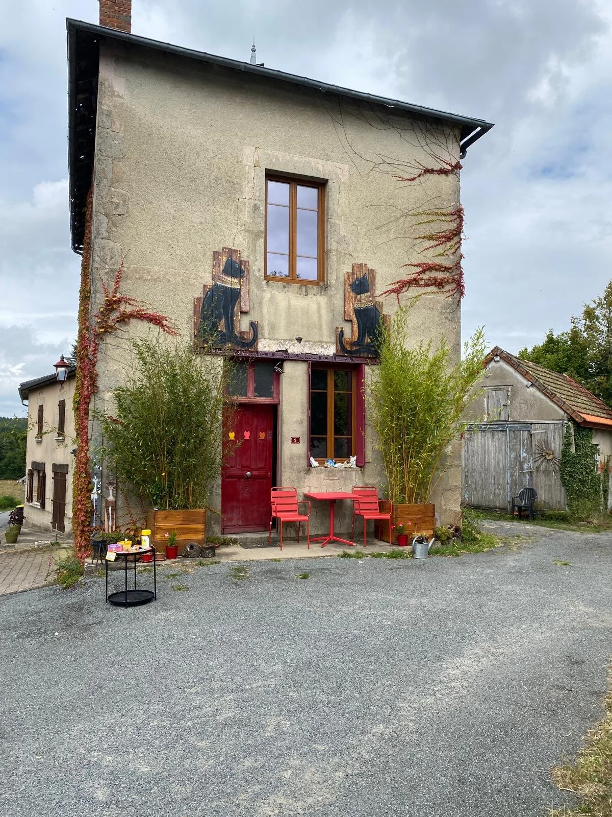 A rustic stone house with Egyptian cat paintings on the front, red door, and outdoor seating area with red chairs, potted plants, and decorative items, set against a cloudy sky.