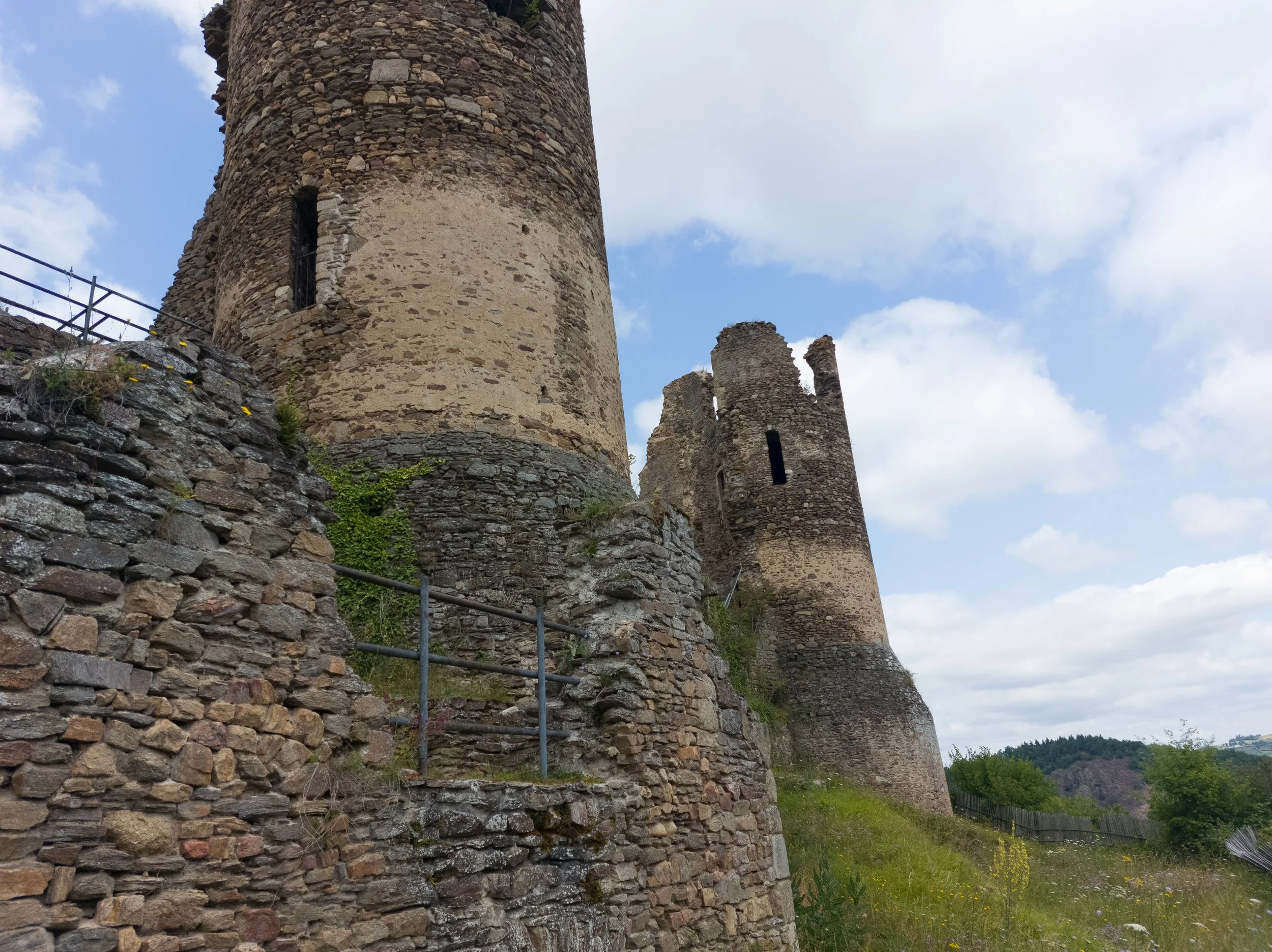 Ancient stone castle ruins with two tall, round towers and a low stone wall in the foreground. Blue sky with scattered clouds in the background.