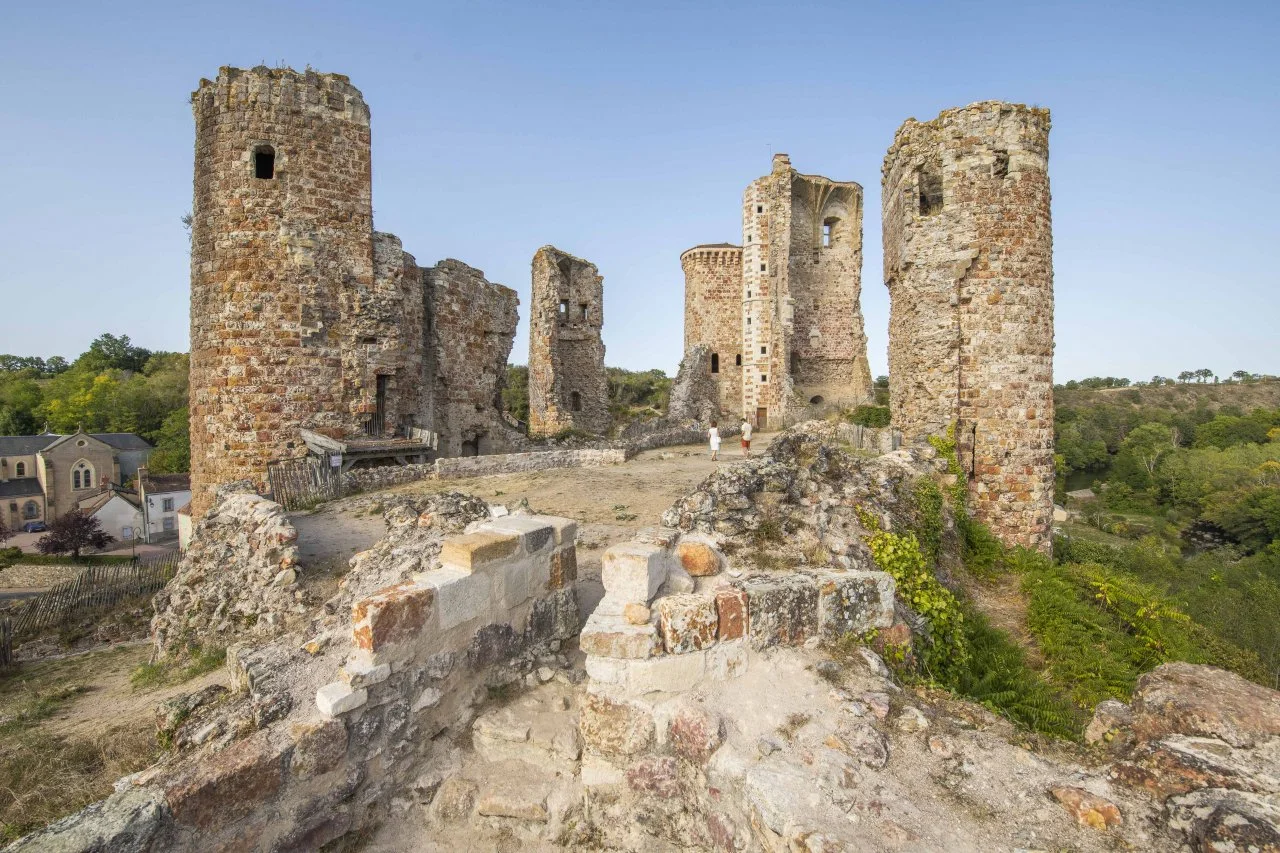 Ruins of an old stone castle with three prominent towers, on a hill with greenery, under a clear blue sky.