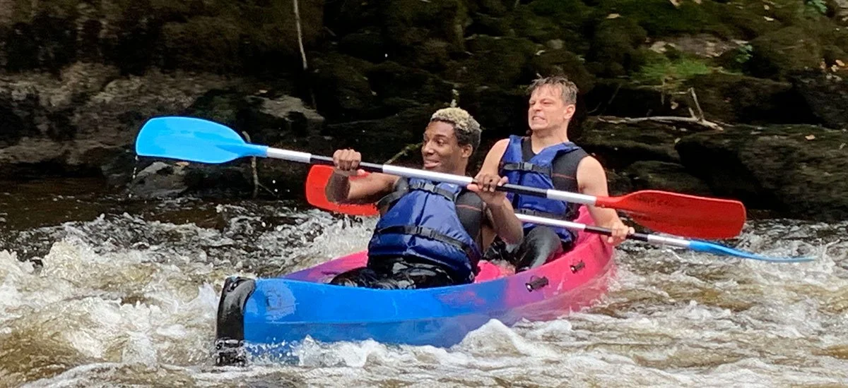 Two people kayaking on a river, paddling through white water. They are wearing life jackets and appear to be enjoying the adventure.