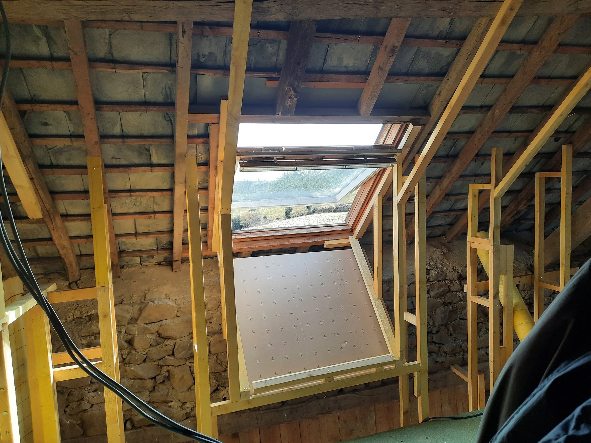 An attic under construction with a skylight window installed in the sloped roof, exposed wooden framing, electrical wiring, and stone wall visible.