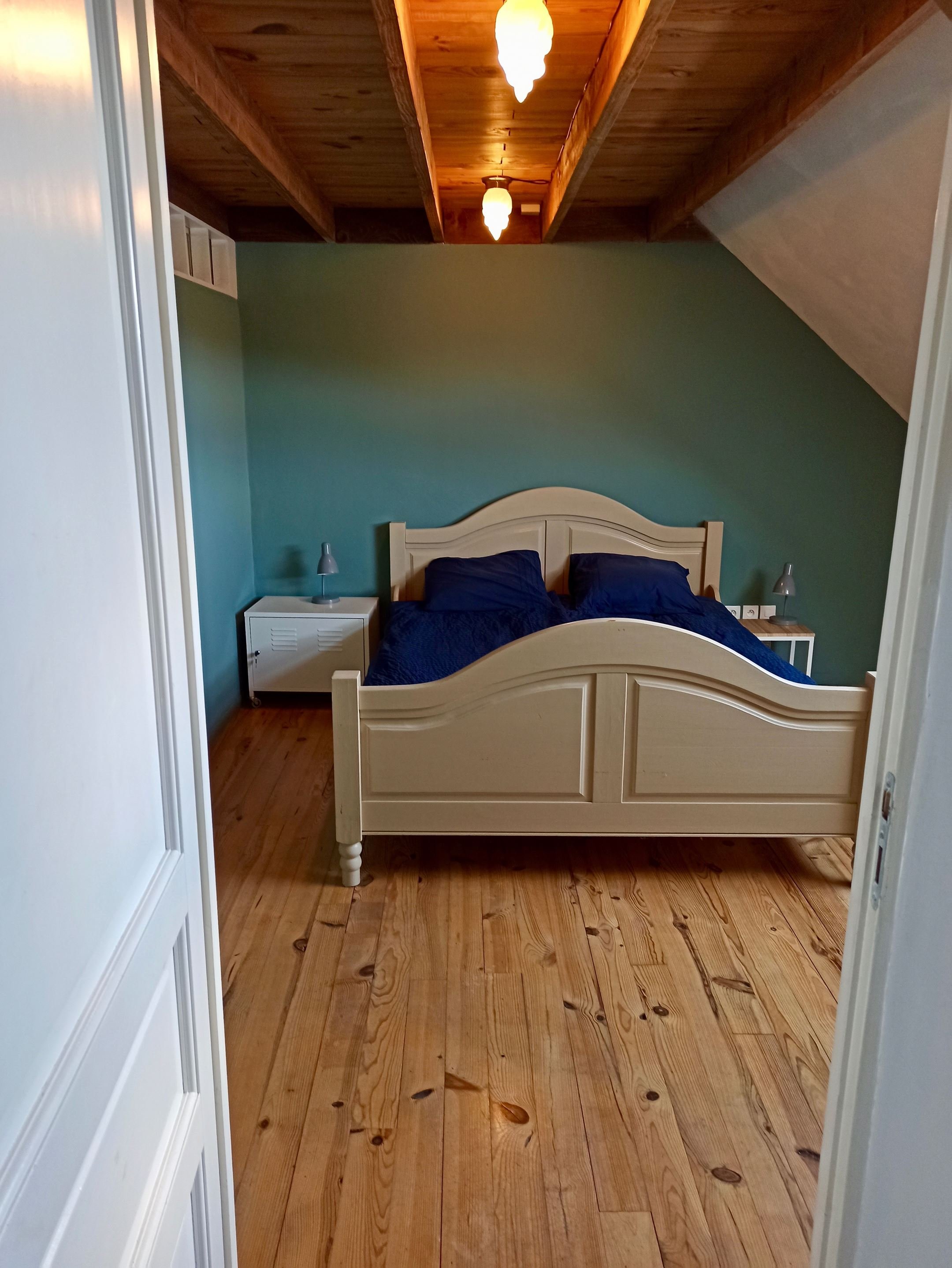 A bedroom with a wooden floor, a cream-colored bed with navy blue bedding, two small white nightstands with matching lamps, and a sloped ceiling with wooden beams and three hanging pendant lights.