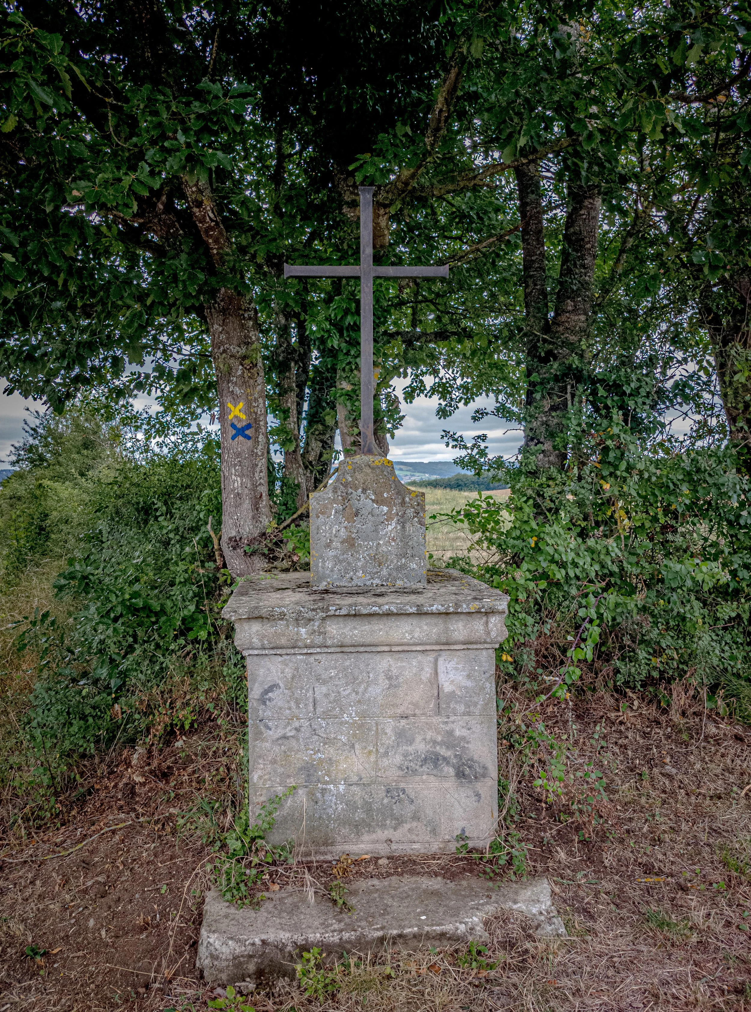 A stone monument with a metal cross on top, situated outdoors among trees and grass.