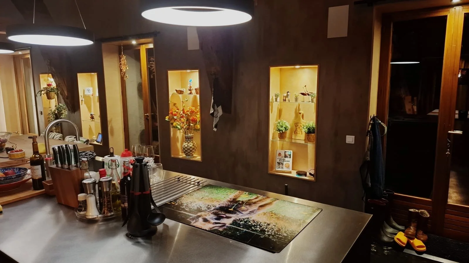 Interior of a kitchen with a counter, various condiments, and utensils. In the background, illuminated wall niches display vases, plants, and decorative items, with hanging pendant lights overhead.