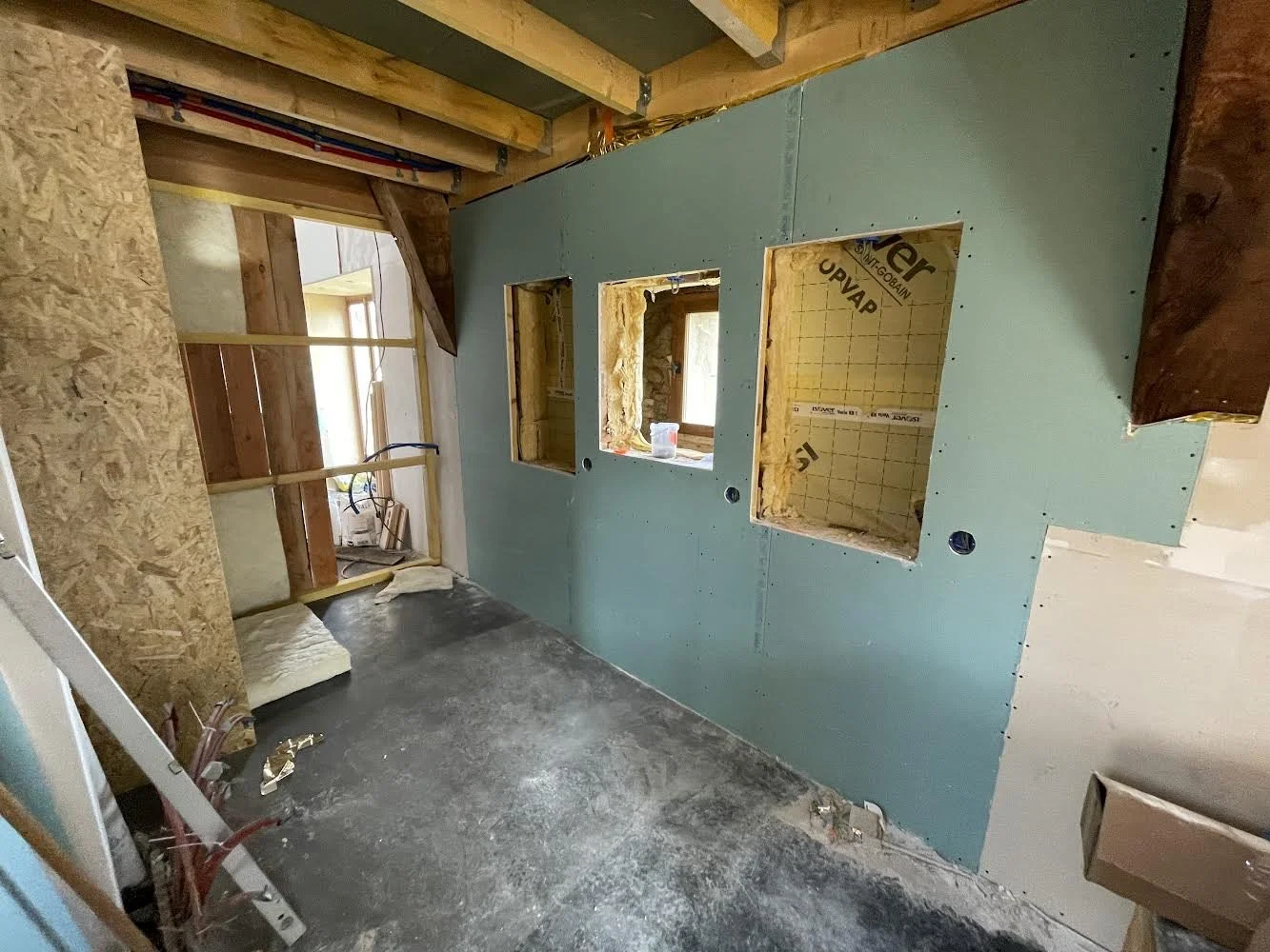 Interior of a construction site showing a partially finished room with drywall and exposed wooden beams, three window openings, and a concrete floor.