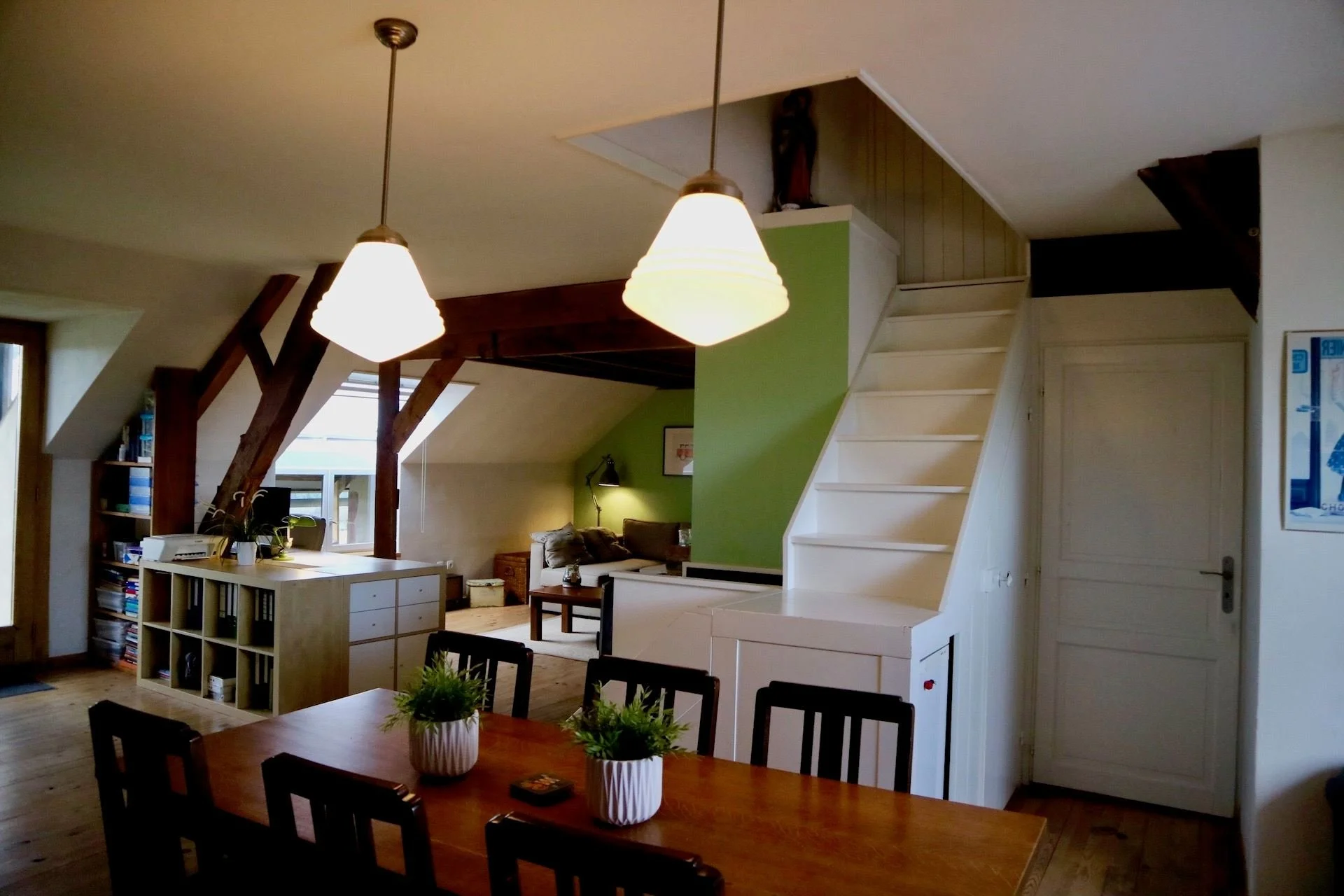 Interior view of a living room and dining area with a staircase, green accent wall, dining table with plants, and seating area with sofa and wall lamp.