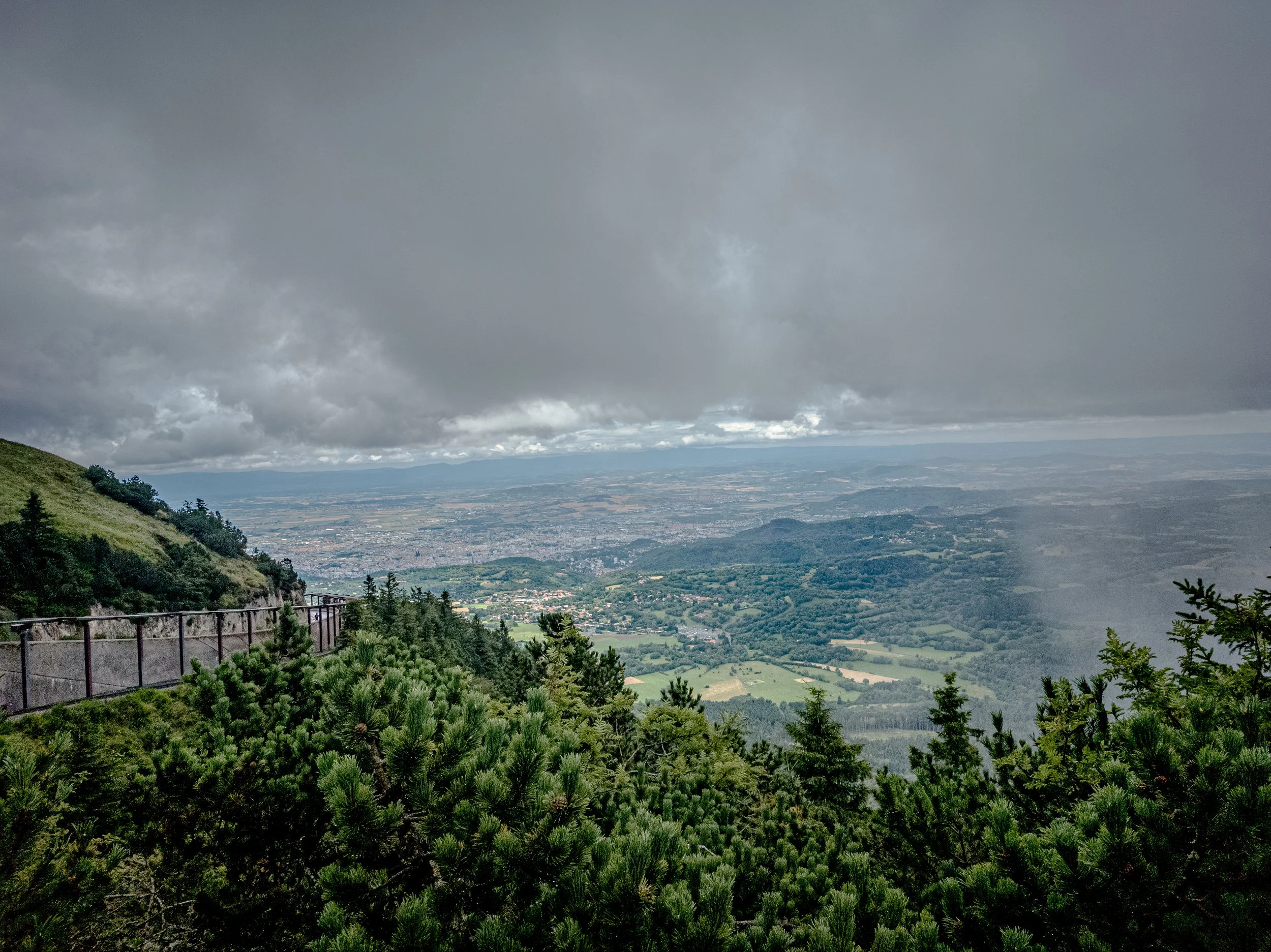 View of a lush green hillside with a metal railing, overlooking a valley with distant city and mountain ranges under a cloudy sky.